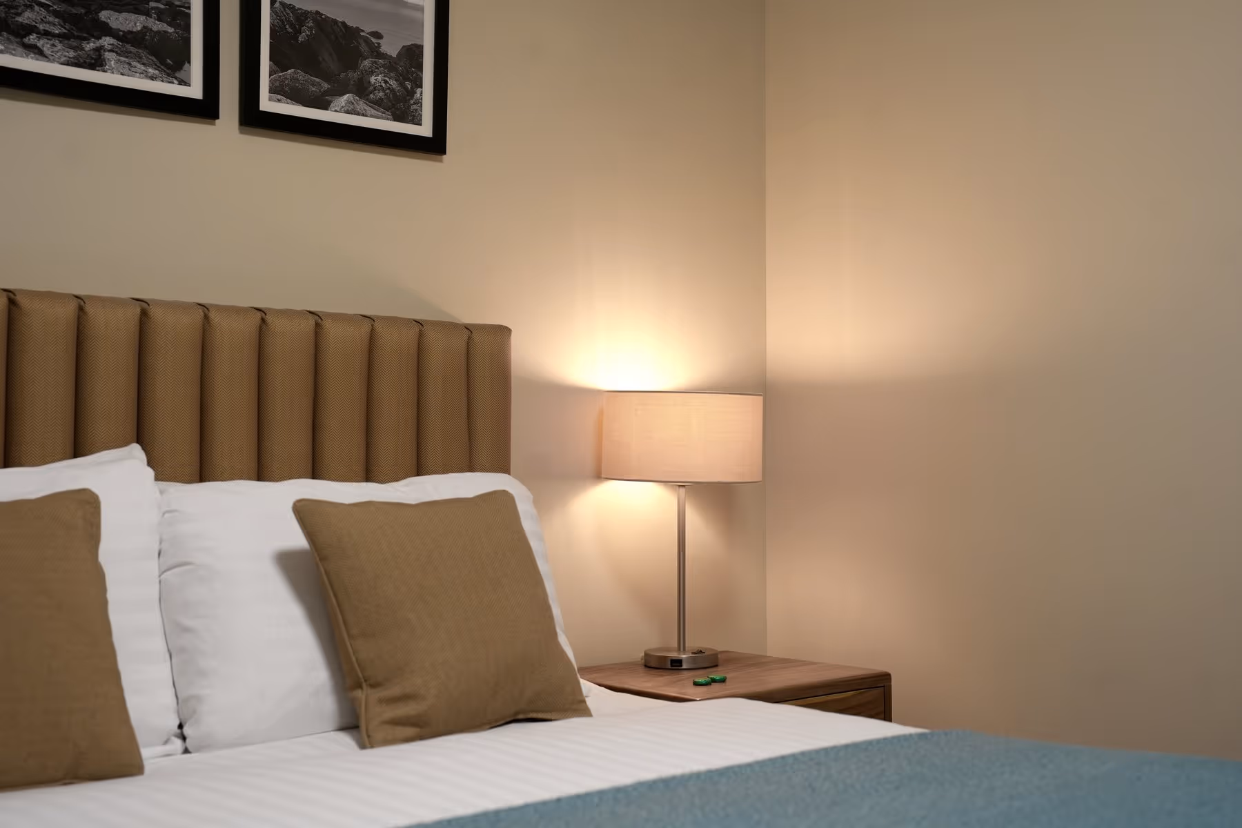 King Room at Riva Helensburgh — close-up of the kingsize bed with a tan channel-stitched headboard, white and tan cushions, and a warm bedside lamp on a walnut nightstand.