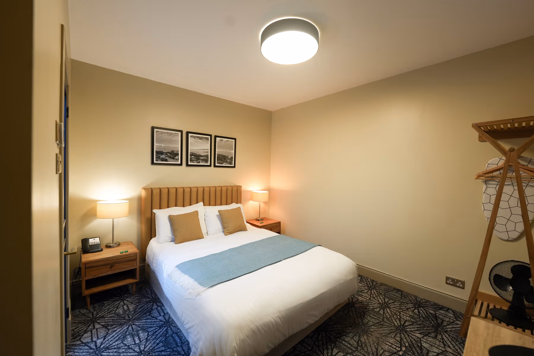 King Room at Riva Helensburgh — overhead view of the kingsize bed with tan headboard, white linen, and coastal prints above, with an en-suite bathroom door visible.
