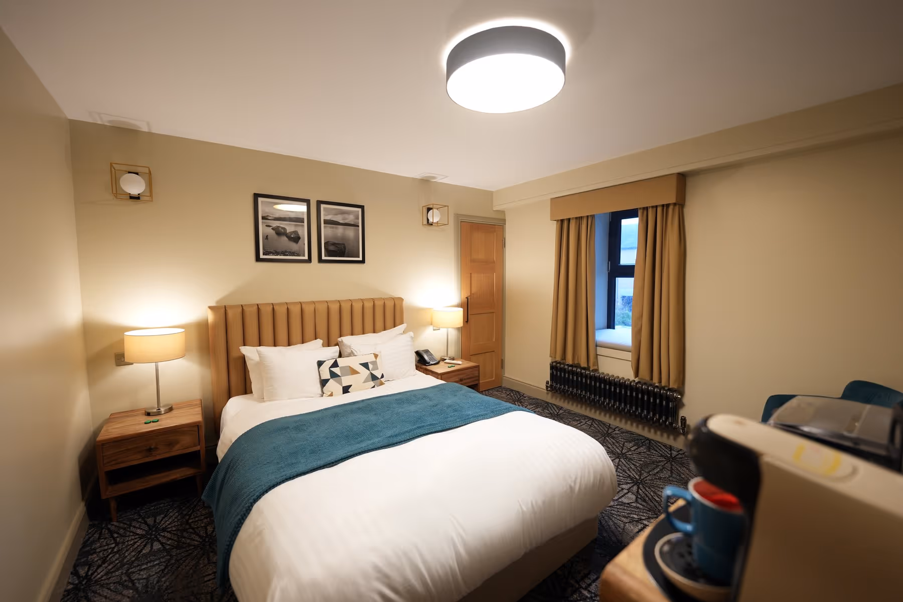 Double Room at Riva Helensburgh — full room view with double bed, tan channel-stitched headboard, teal throw, wall sconces, and a tray with espresso machine in the foreground.