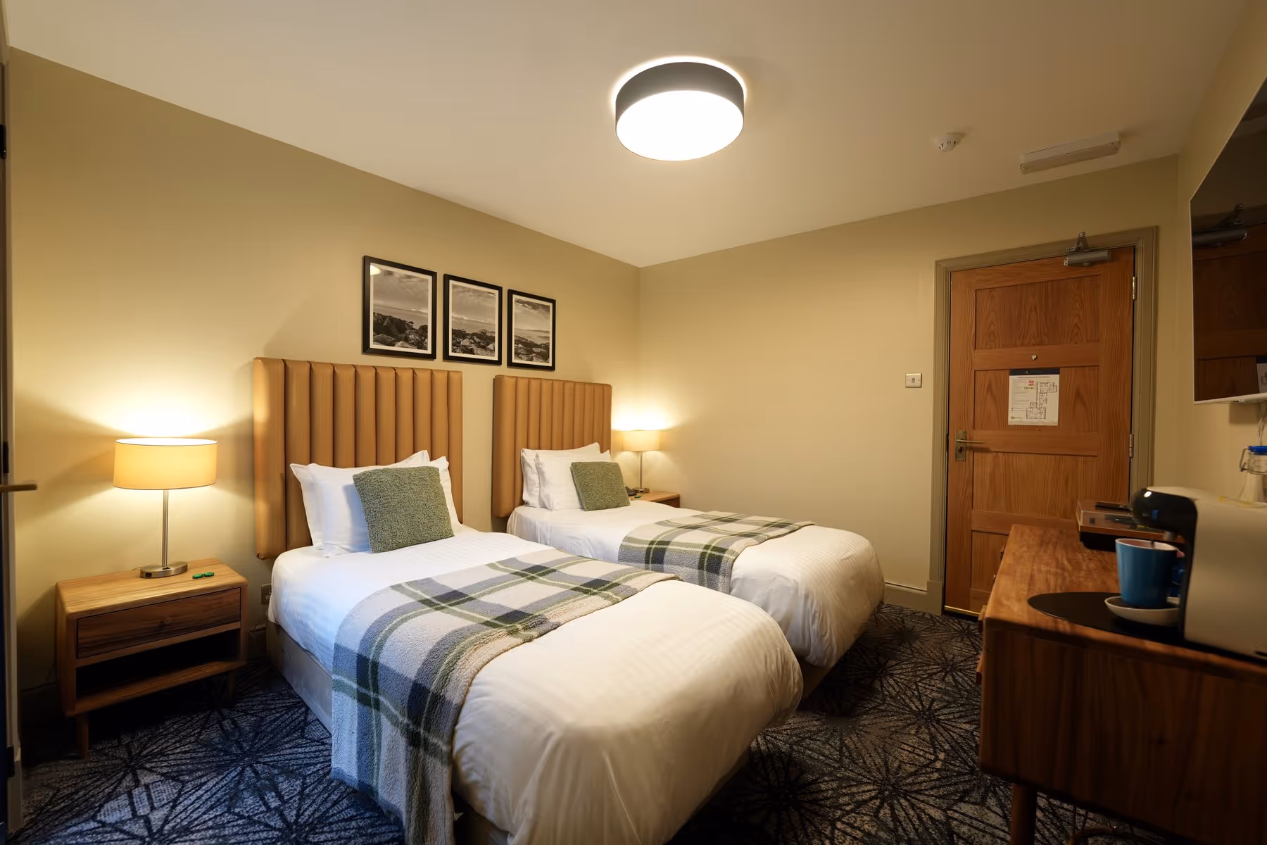 Twin Room at Riva Helensburgh — full room view with two single beds, tan upholstered headboards, green tartan throws, a shared bedside lamp, and a work desk with espresso machine.