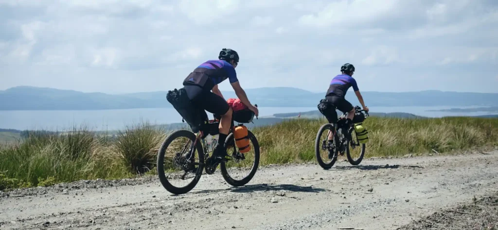 Two cyclists on a gravel track above the Firth of Clyde near Helensburgh, with sweeping views over the water and distant hills on a bright day.