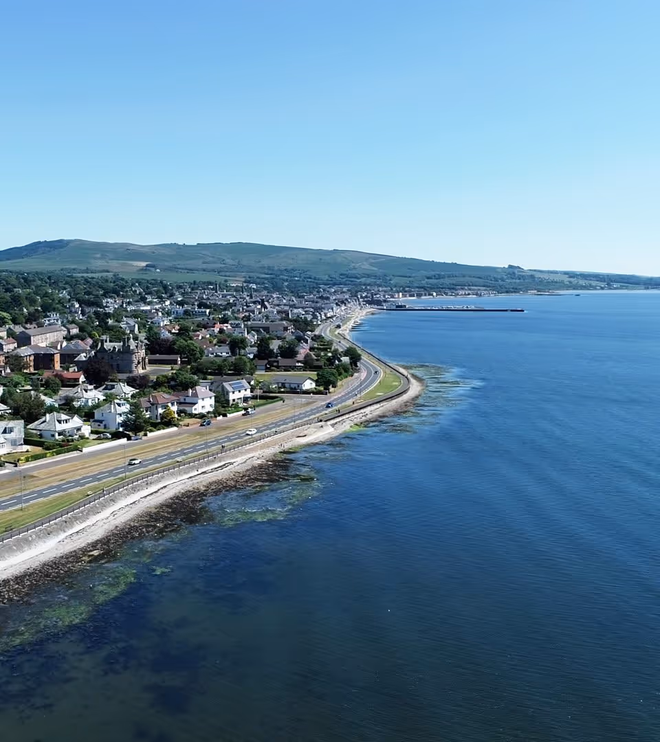 Aerial view of Helensburgh seafront showing the coastal road running alongside the Firth of Clyde, with the town and hills of Argyll in the background.