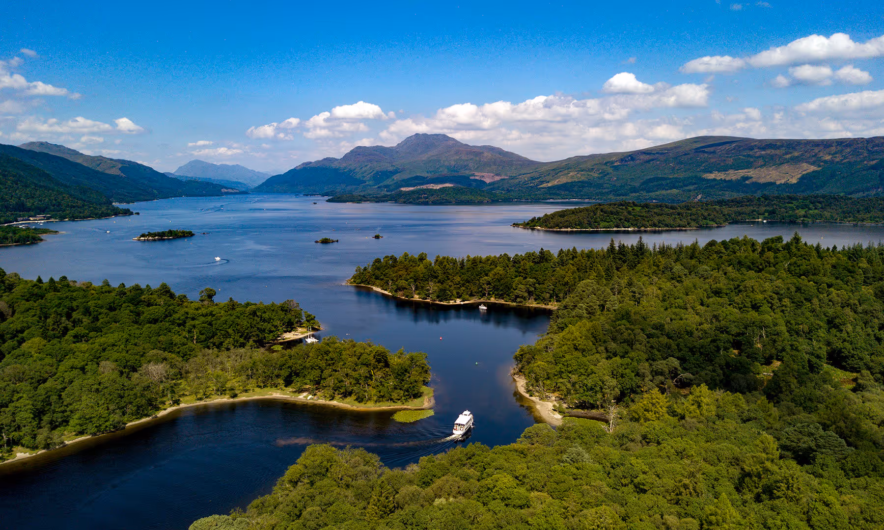 Aerial view of Loch Lomond, Scotland, with wooded islands, a boat navigating a narrow channel, and Ben Lomond rising in the background under a blue sky.