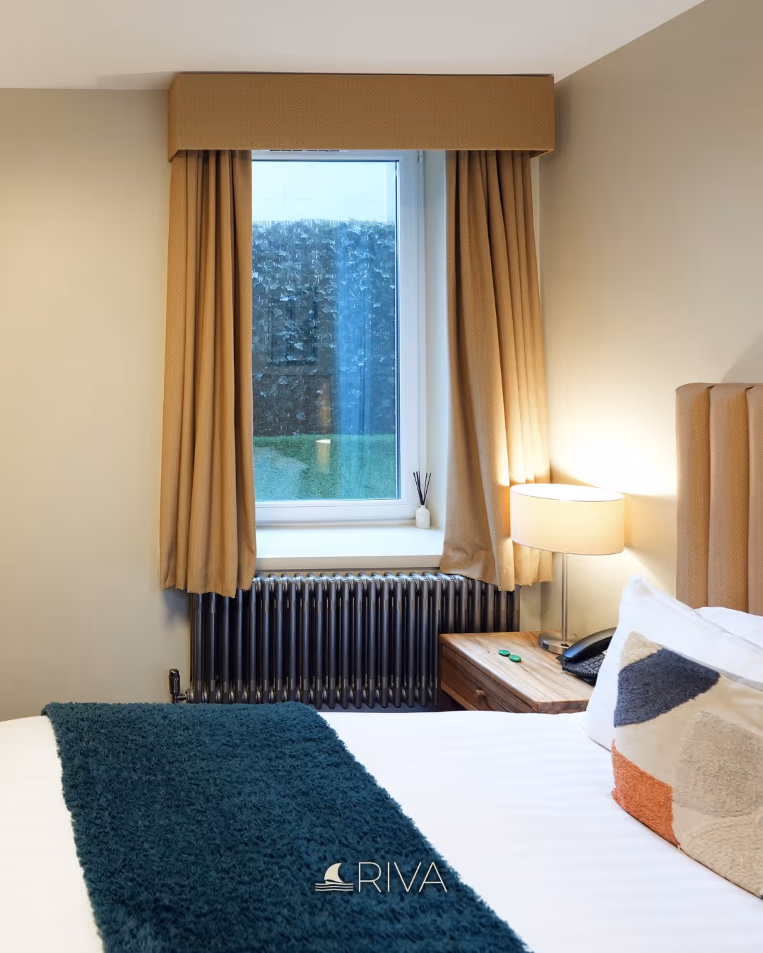 Cozy bedroom window with beige curtains, radiator, bedside table with lamp and patterned cushion on bed.