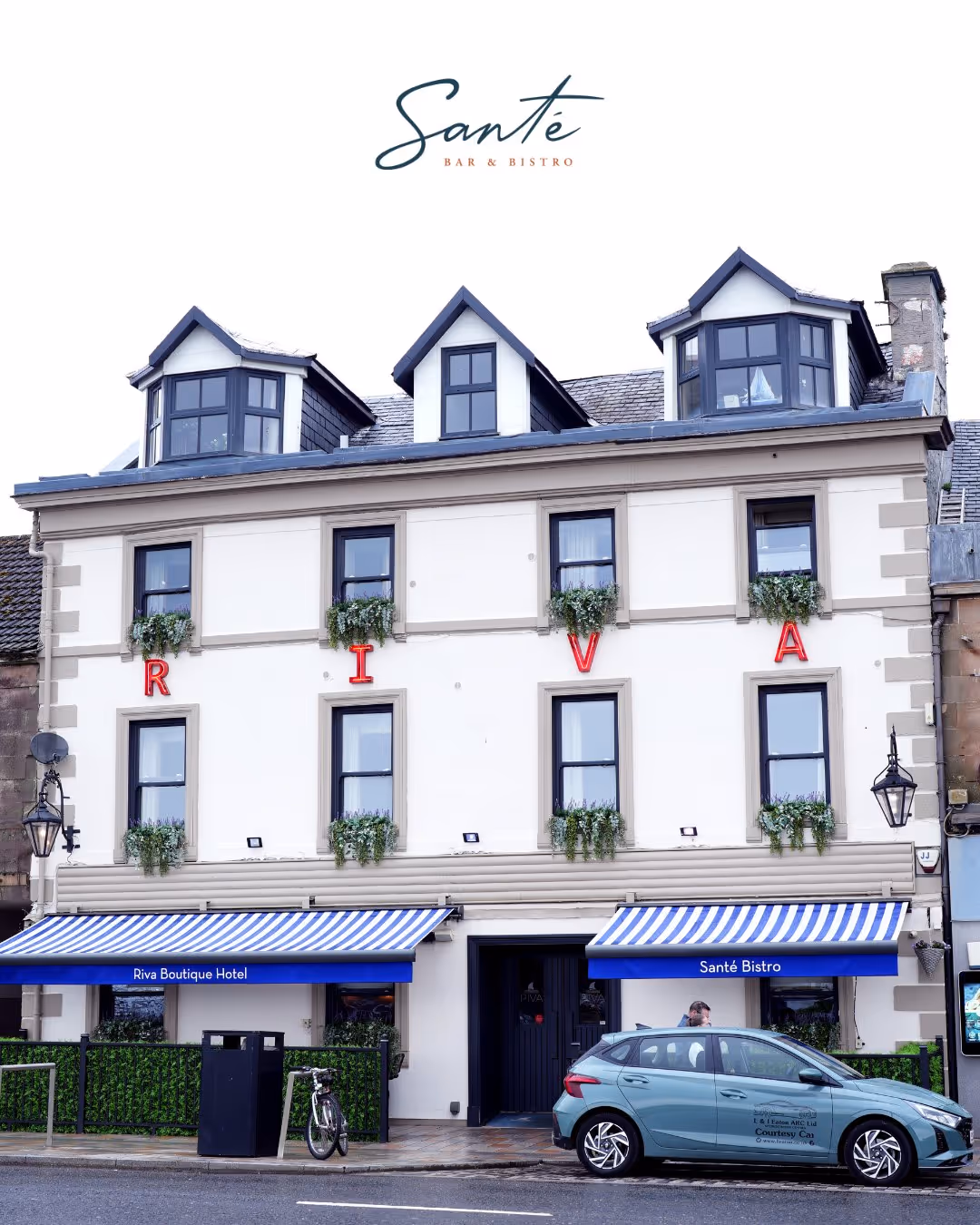 Front facade of Riva Boutique Hotel and Santé Bistro with striped awnings and a parked car.