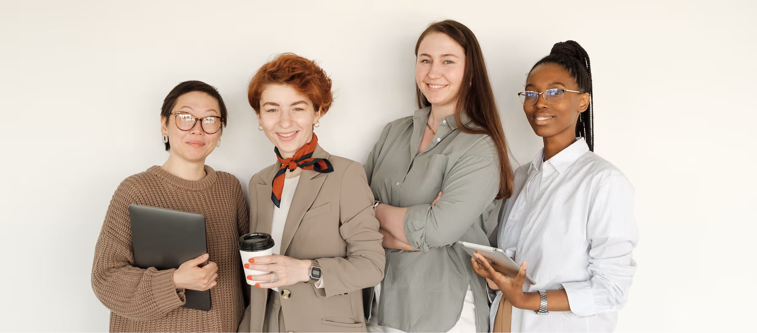 Four diverse women smiling, one holding a laptop, another a coffee cup, one with arms crossed, and one a tablet.