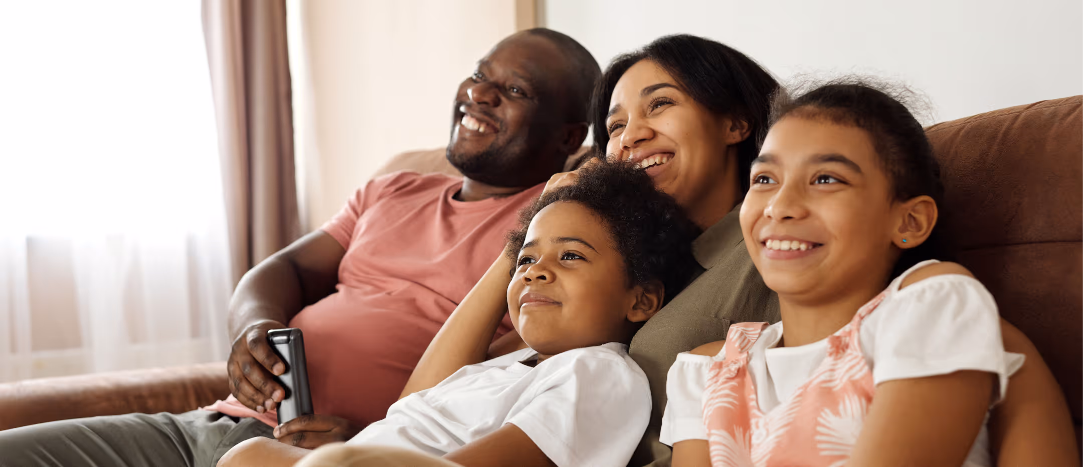 Smiling family of four sitting on a couch watching TV together in a cozy living room.