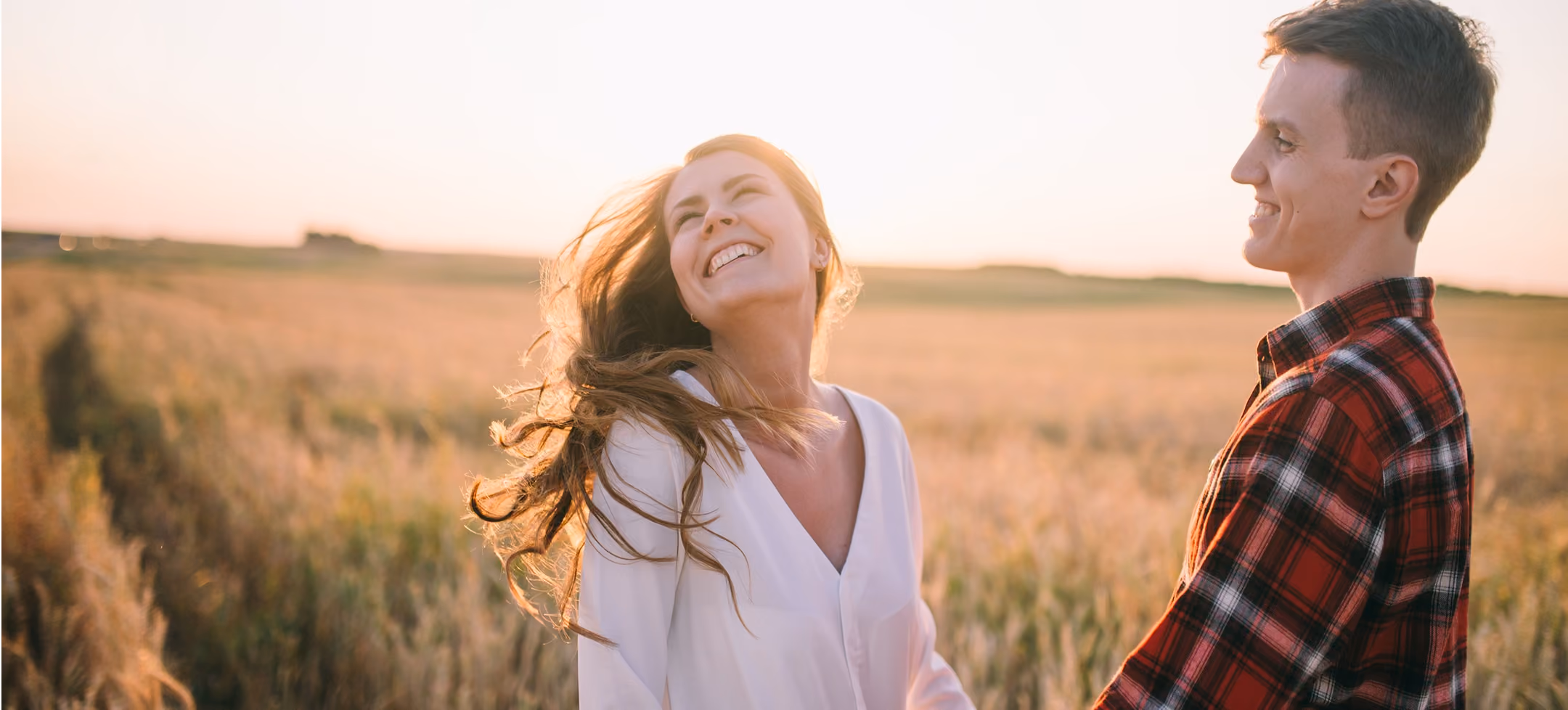 Smiling couple holding hands in a sunlit field at sunset, woman in white blouse, man in red plaid shirt.