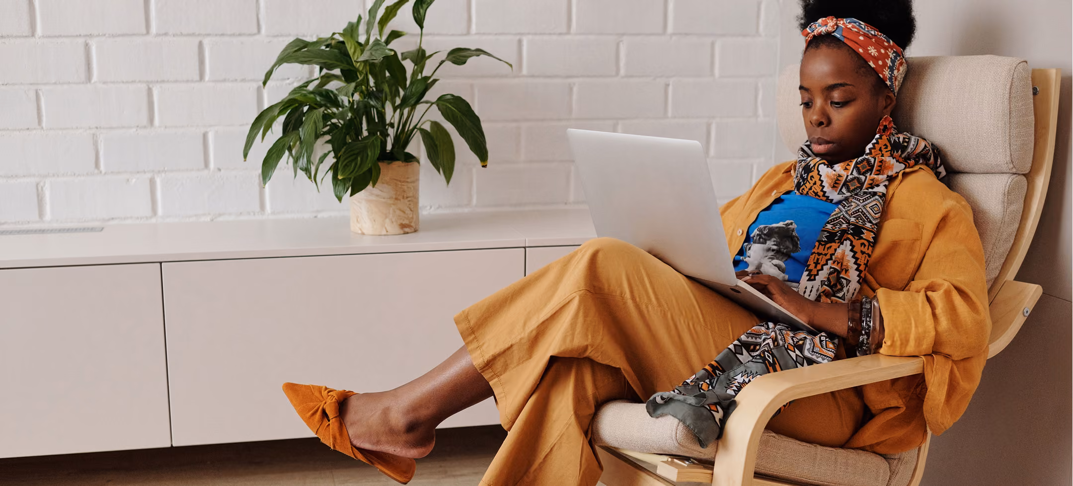 Woman in patterned scarf and orange outfit sitting in a chair using a laptop near a white cabinet with a plant.
