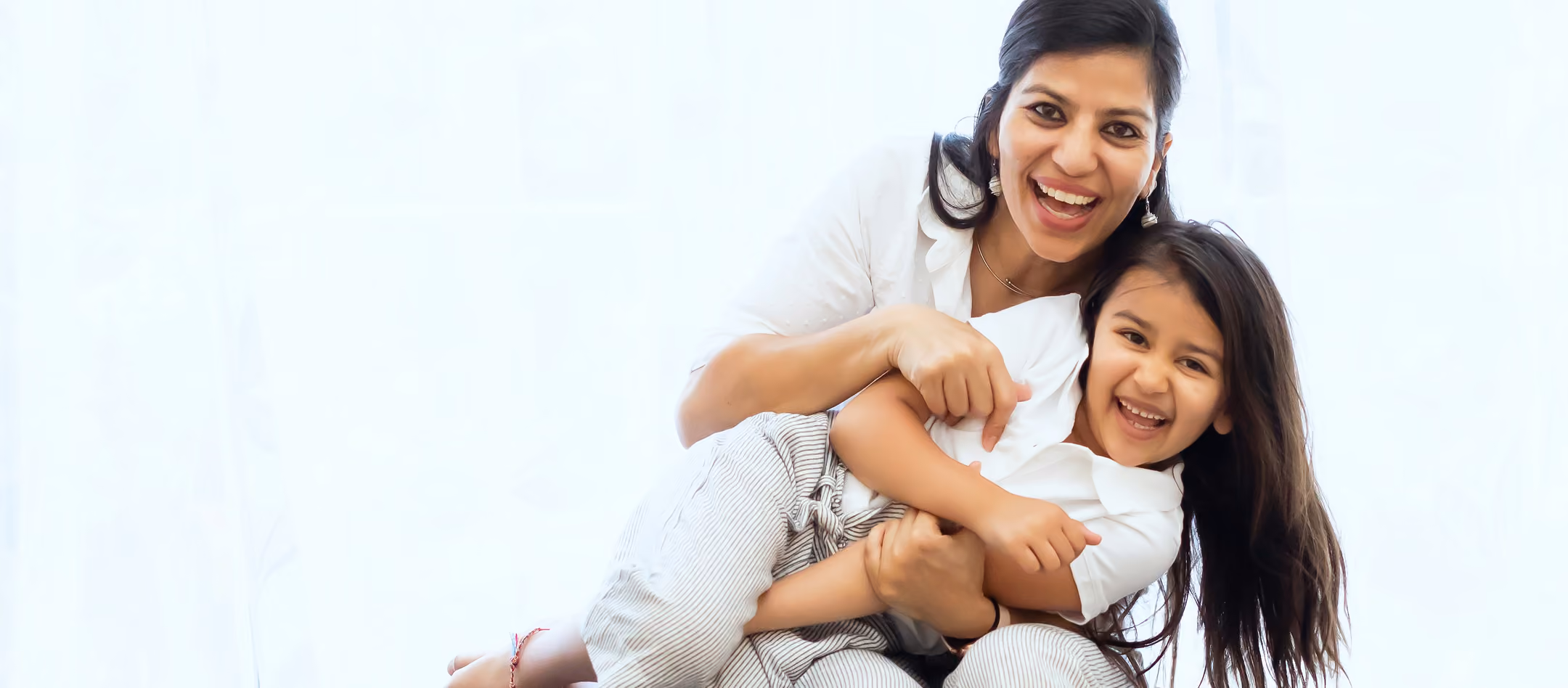 Smiling woman hugging a laughing young girl with long hair against a white background.