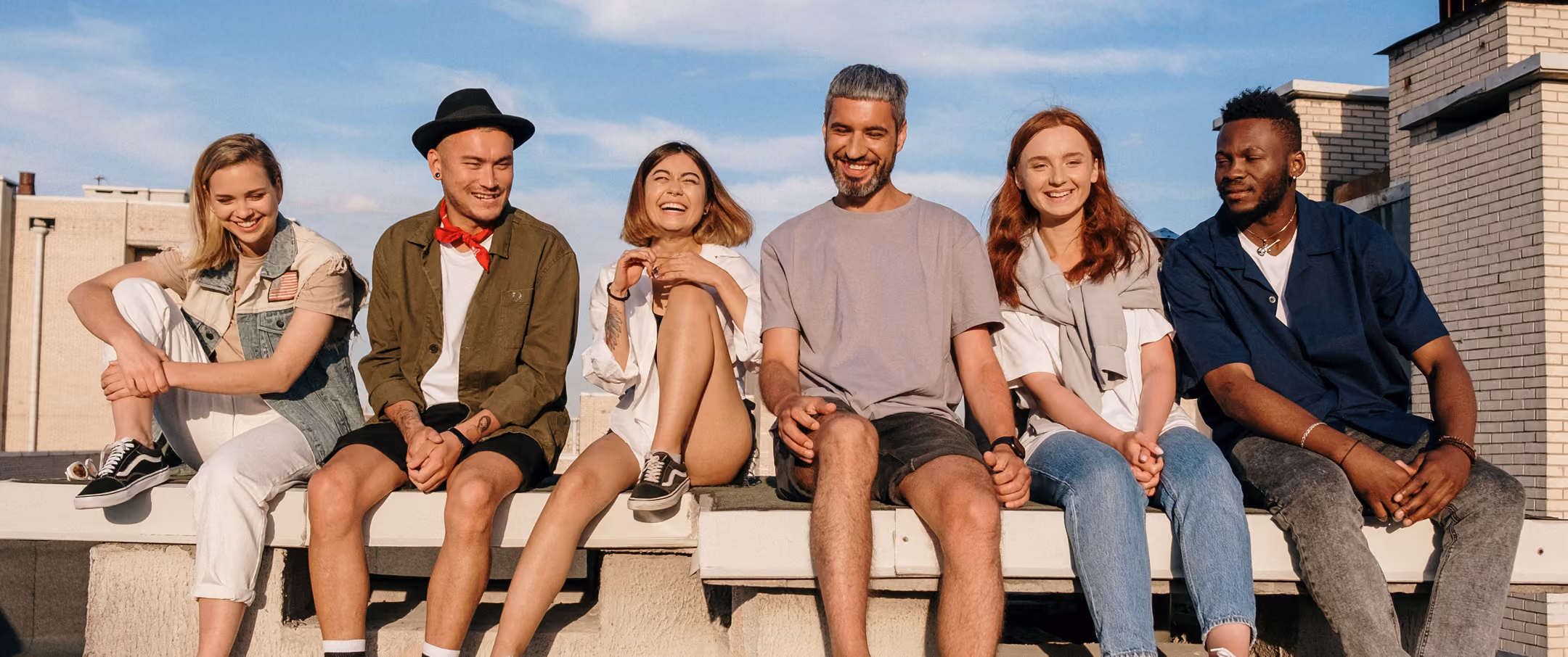 Six diverse young adults sitting on a rooftop ledge, smiling and enjoying sunny weather.