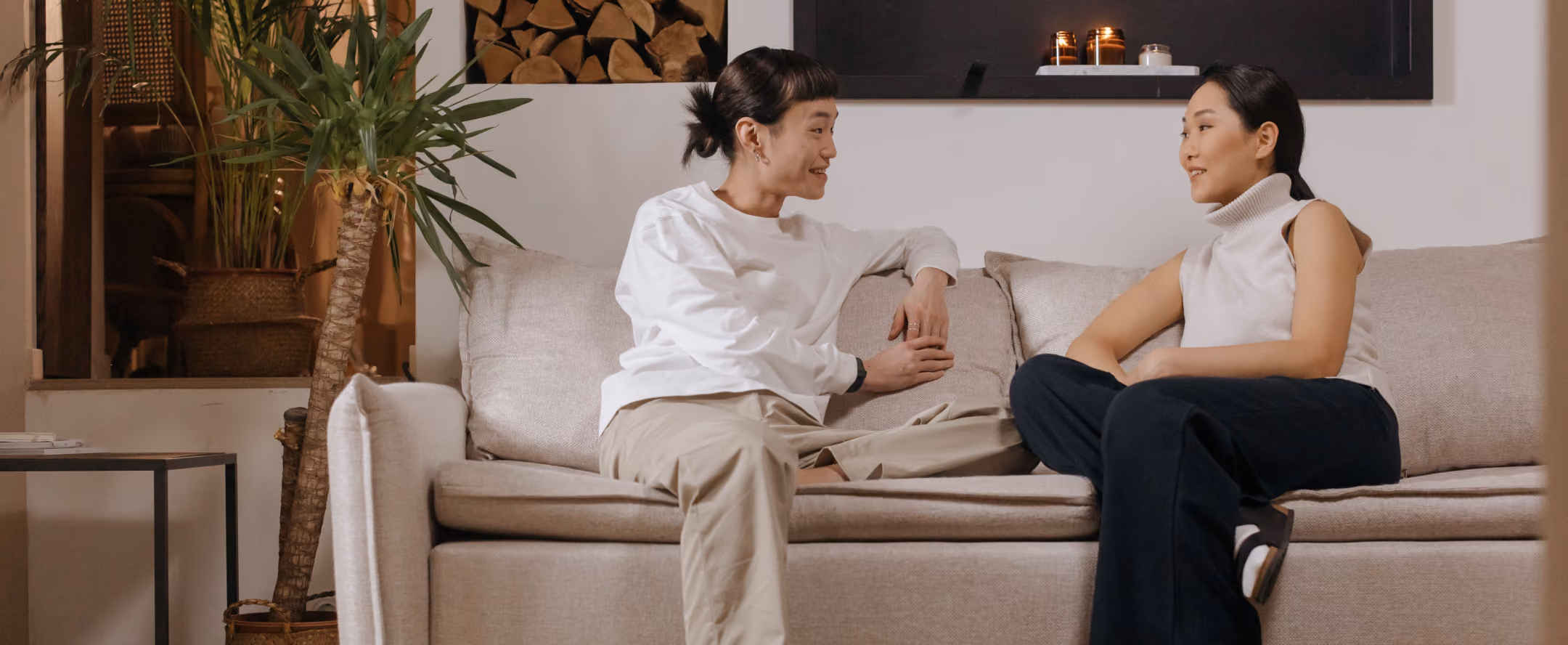 Two women sitting on a beige sofa facing each other and engaged in a conversation in a cozy living room with plants and candles in the background.