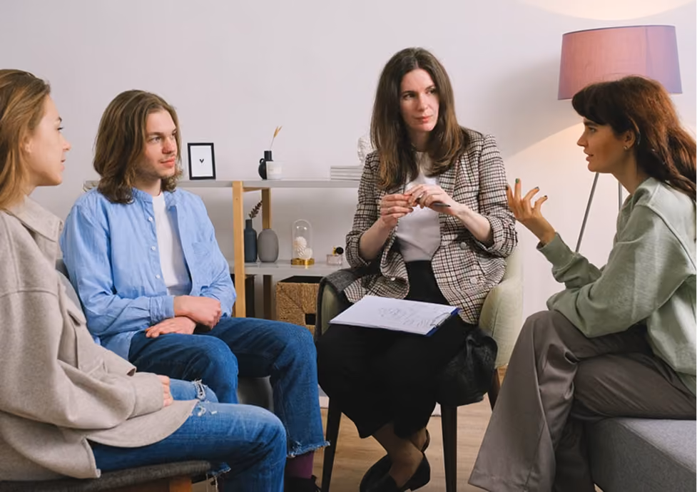 Four people sitting in a circle in a living room having a conversation, including a woman holding papers, suggesting a group therapy or counseling session.