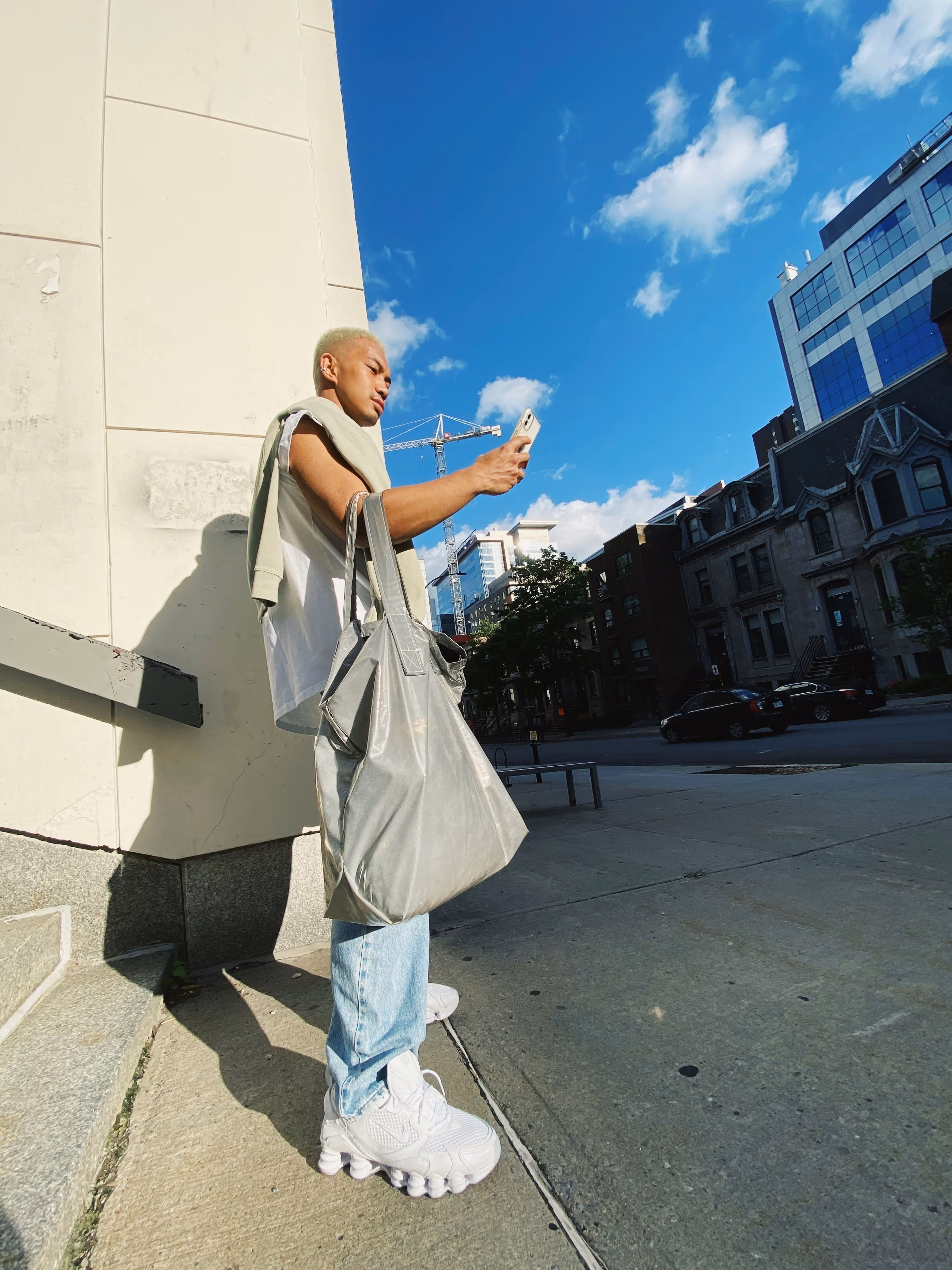 Person standing on a city sidewalk wearing light jeans and white sneakers, holding a phone and large bag.