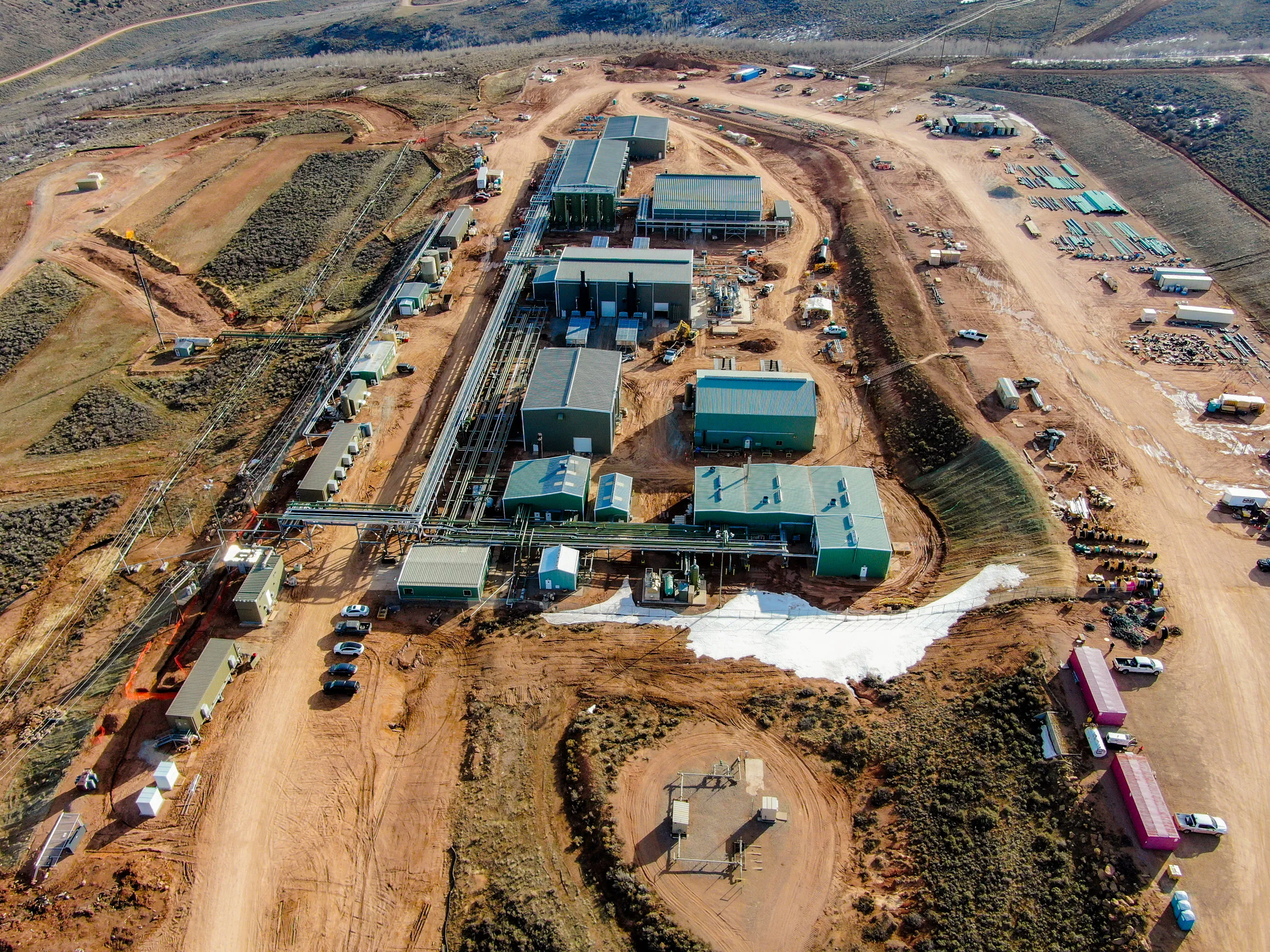 Aerial view of an industrial facility with multiple green buildings, pipes, and dirt roads in a rural area.
