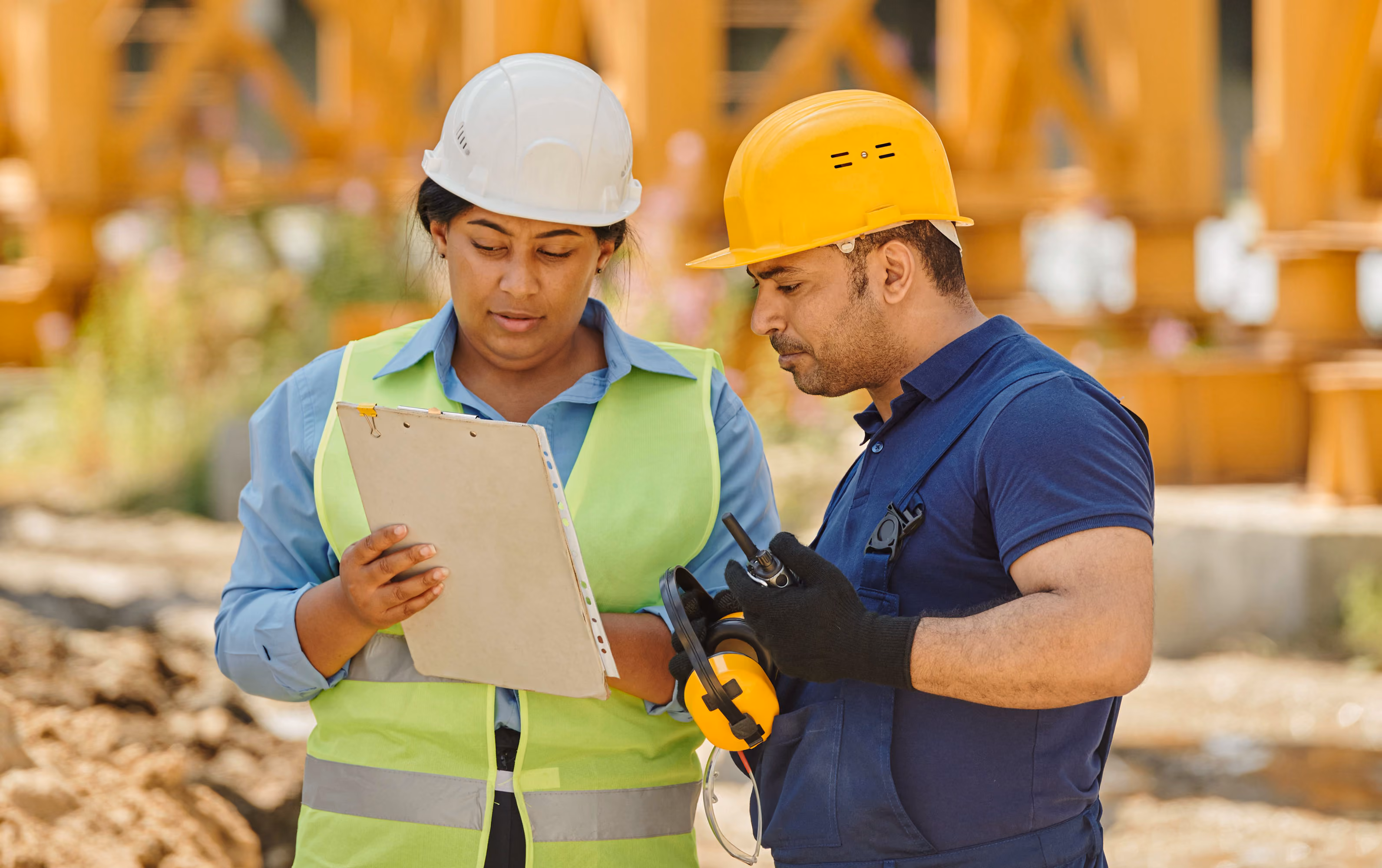Two construction workers wearing helmets discuss plans on a clipboard at a work site.