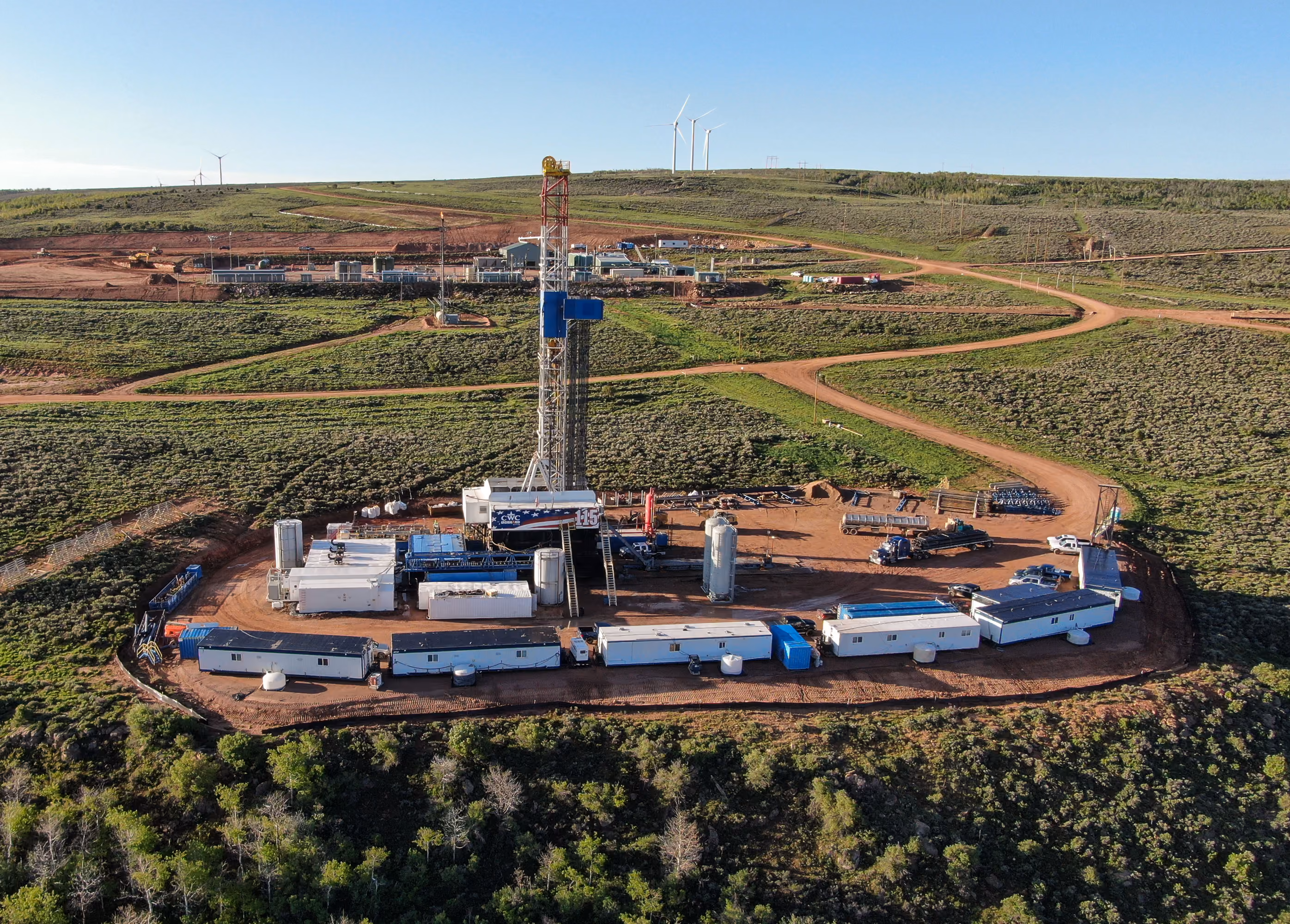 Aerial view of an oil drilling rig surrounded by equipment and trailers in a green, open landscape.