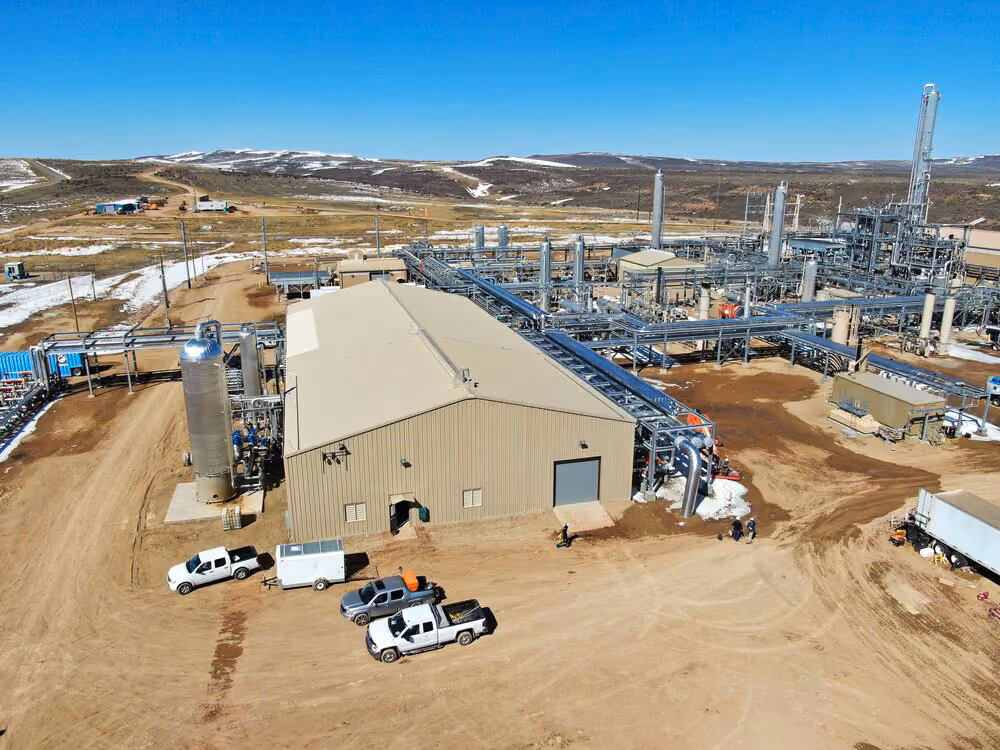 Industrial plant with beige building, pipes, and trucks on a dirt road in a rural area with snow patches.