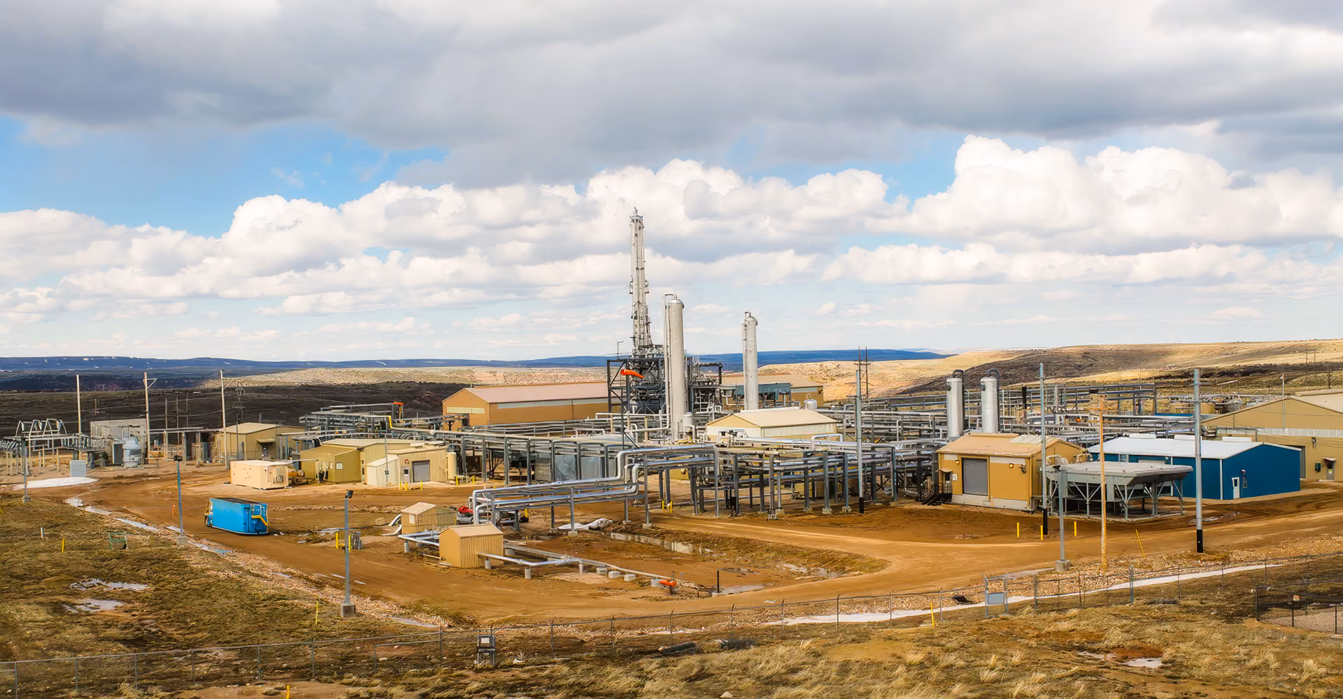 Wide view of an industrial plant with pipes, yellow buildings, and cloudy sky in a rural area.