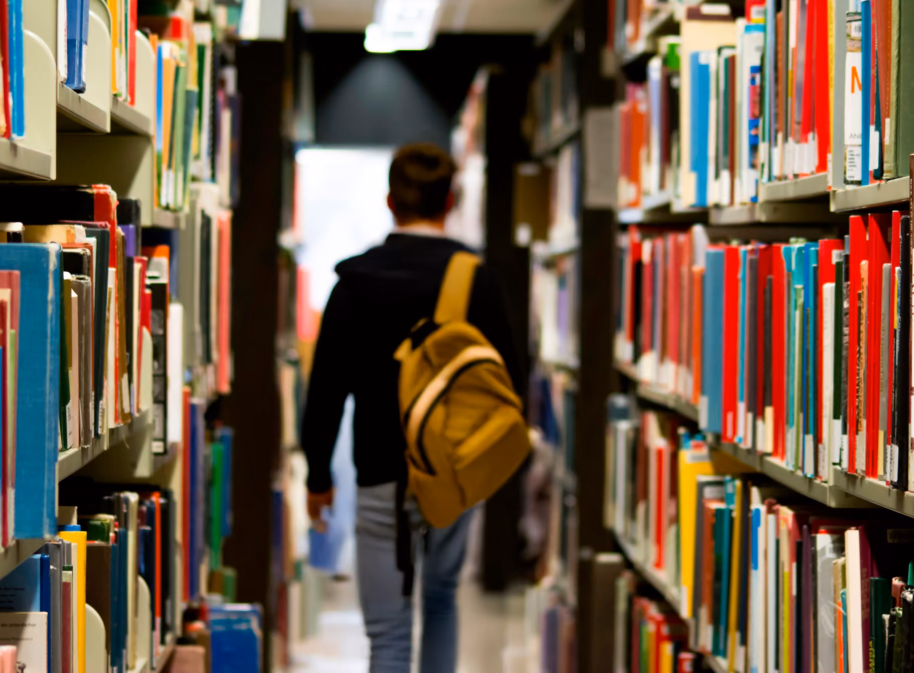 Person with yellow backpack walking between bookshelves in a library aisle.
