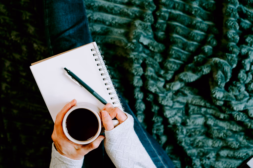 Personne tenant une tasse de café avec un cahier blanc et un stylo sur les genoux, assise sur un tapis vert.