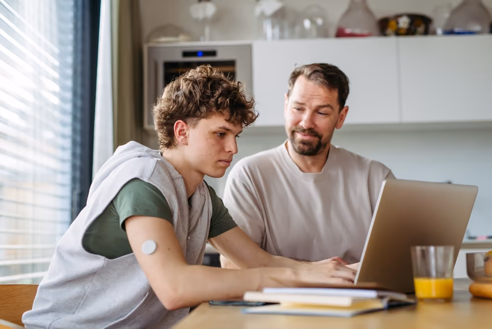 Teen boy and man sitting at table using a laptop in a bright kitchen with a glass of orange juice nearby.