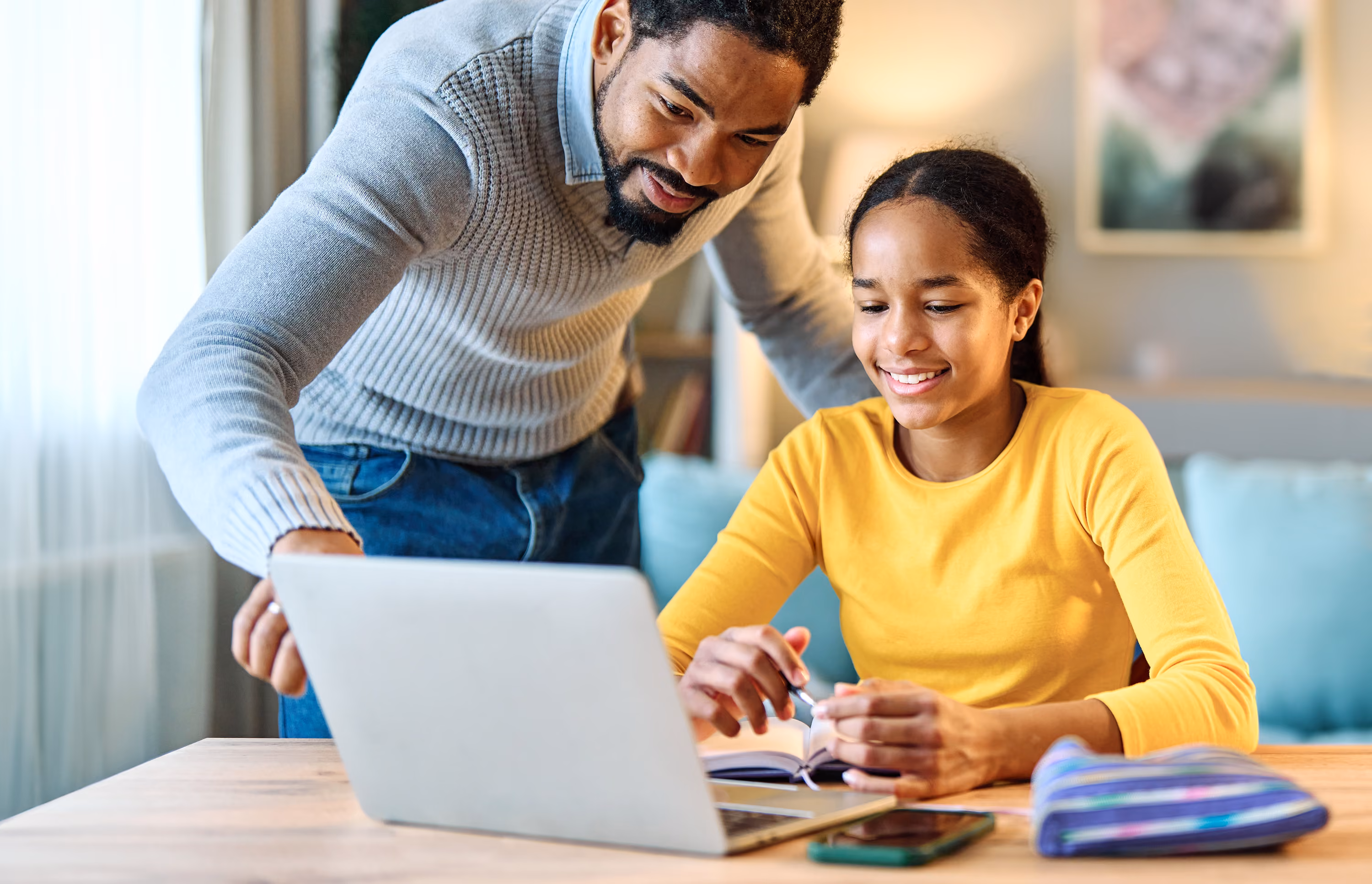 Smiling man helps girl with homework as they look at a laptop together at home.