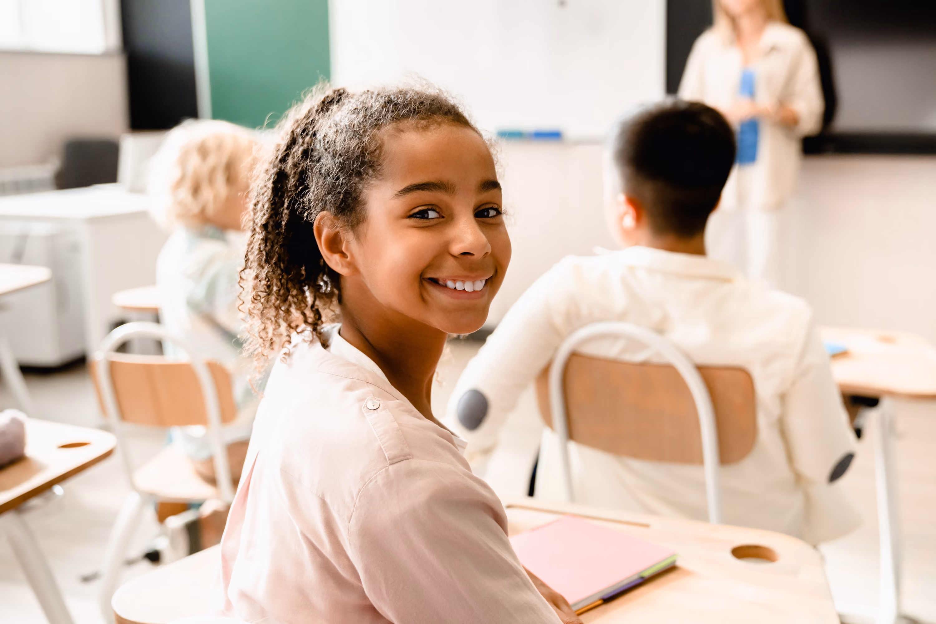 Smiling girl sitting at desk in classroom with other students and teacher in the background.