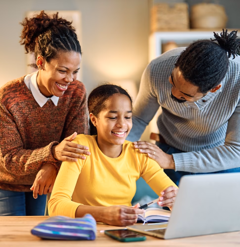 Smiling parents support their daughter working on a laptop and notebook at a wooden table.