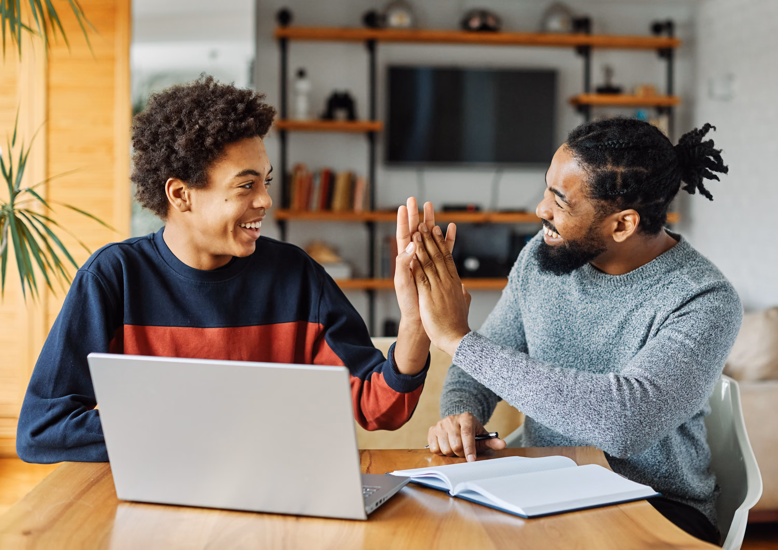 Two smiling men sitting at a table with a laptop and book, giving each other a high five.