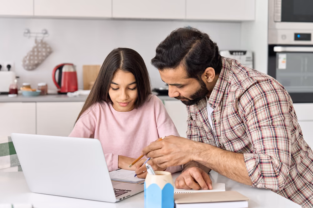 Man helping girl with homework at a kitchen table with a laptop and notebooks.