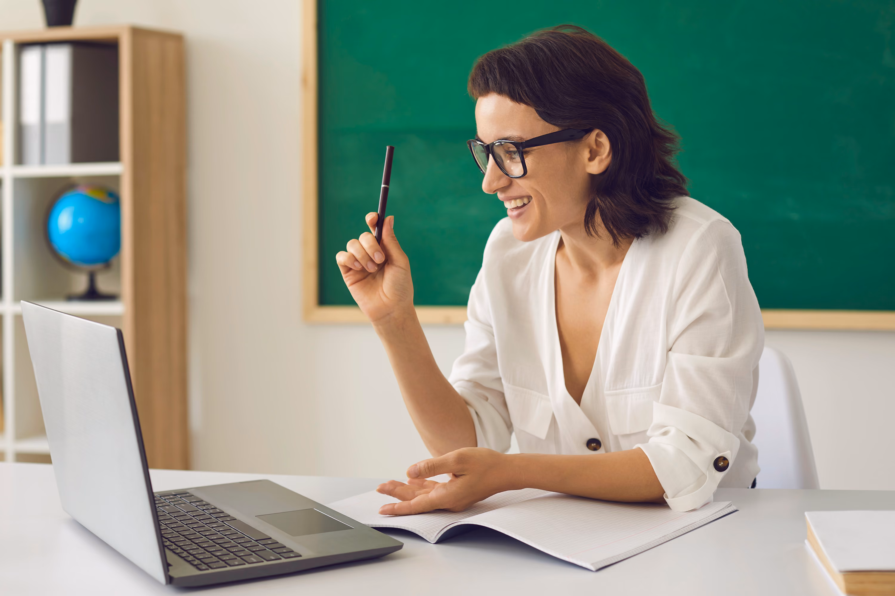 Woman with glasses sitting at a desk smiling while holding a pen and looking at a laptop screen.