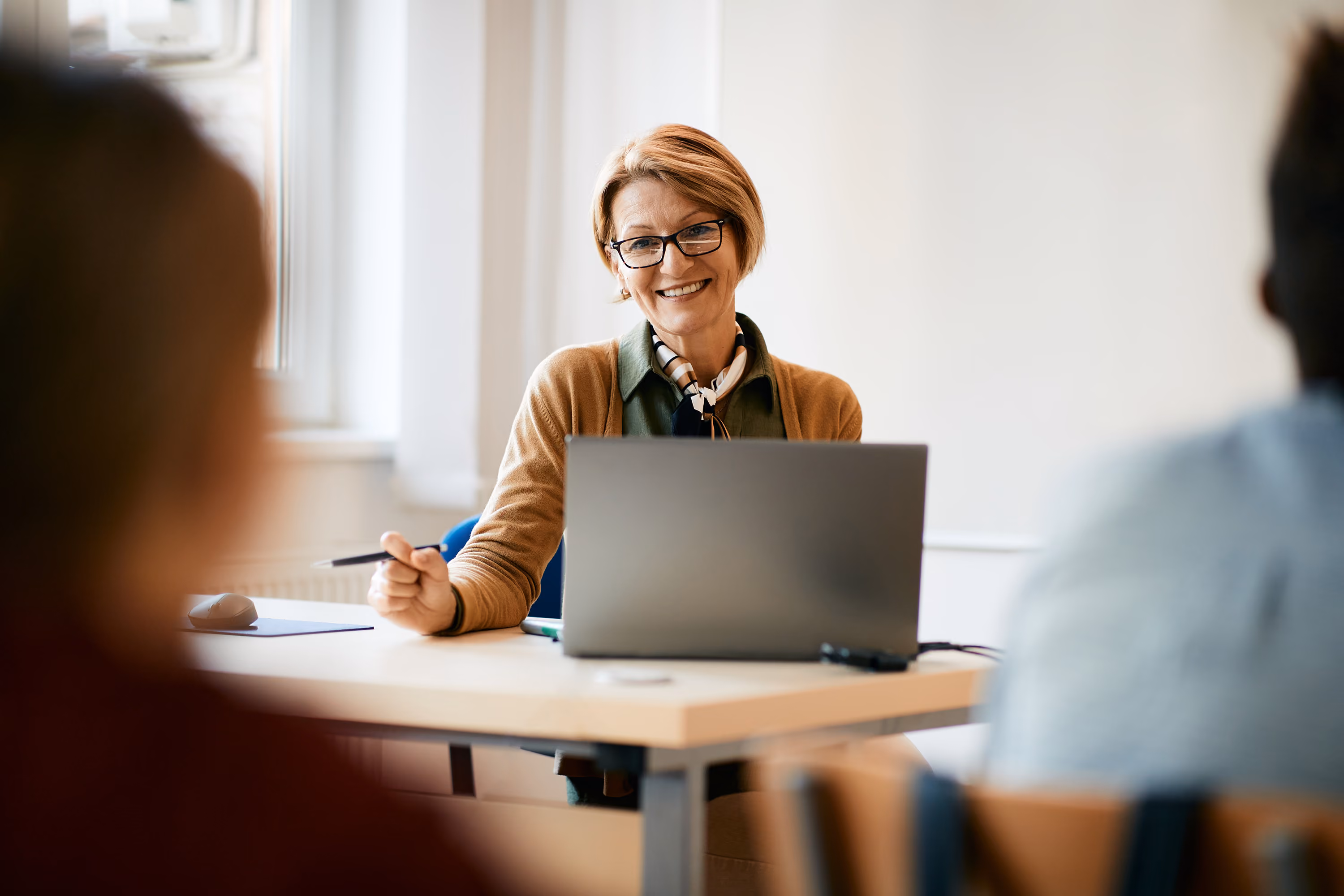 Smiling woman wearing glasses and a scarf, sitting at desk with laptop and holding a pen in a meeting.