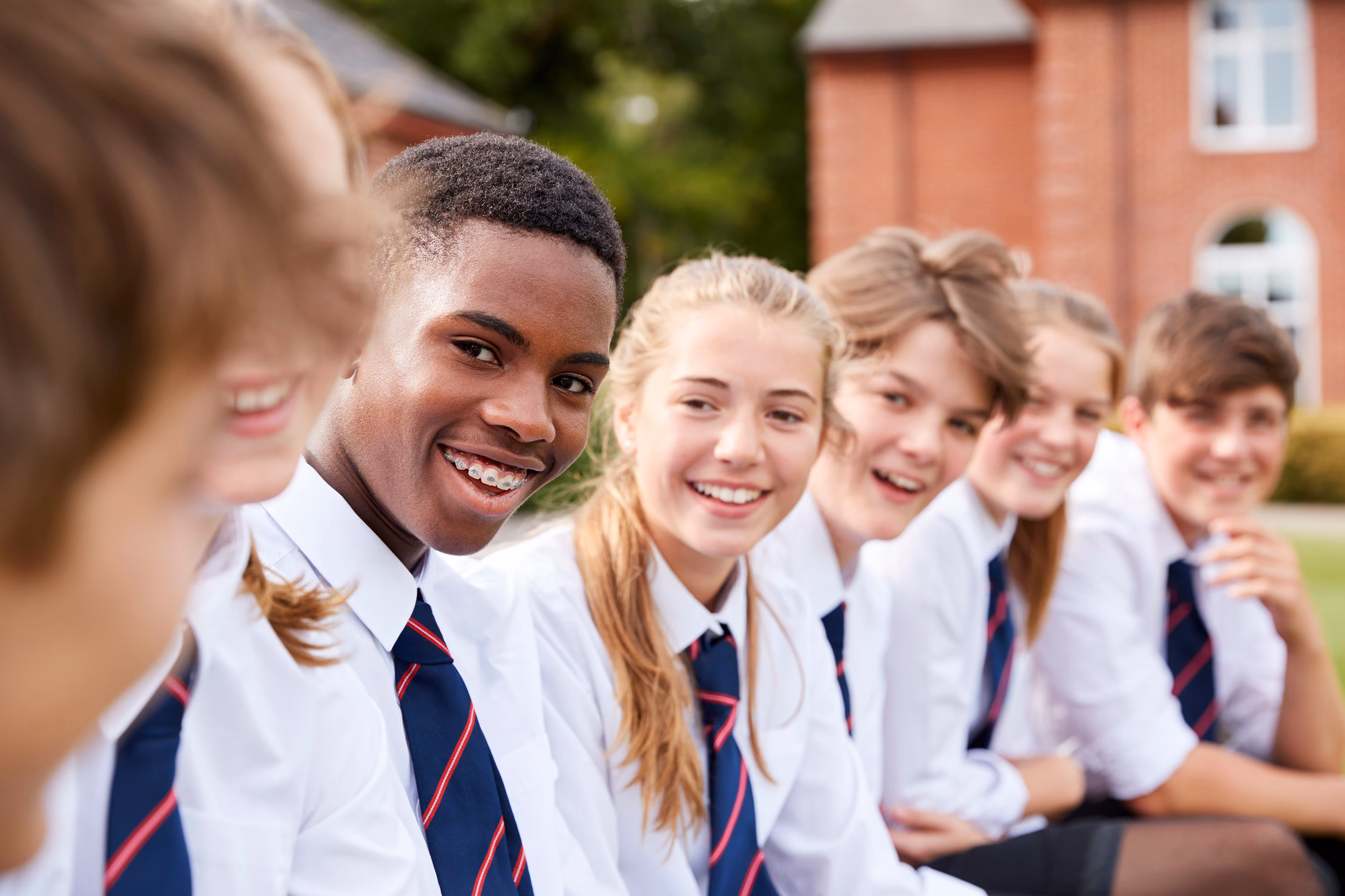 Group of six smiling high school students in uniforms sitting outside near a red brick building.