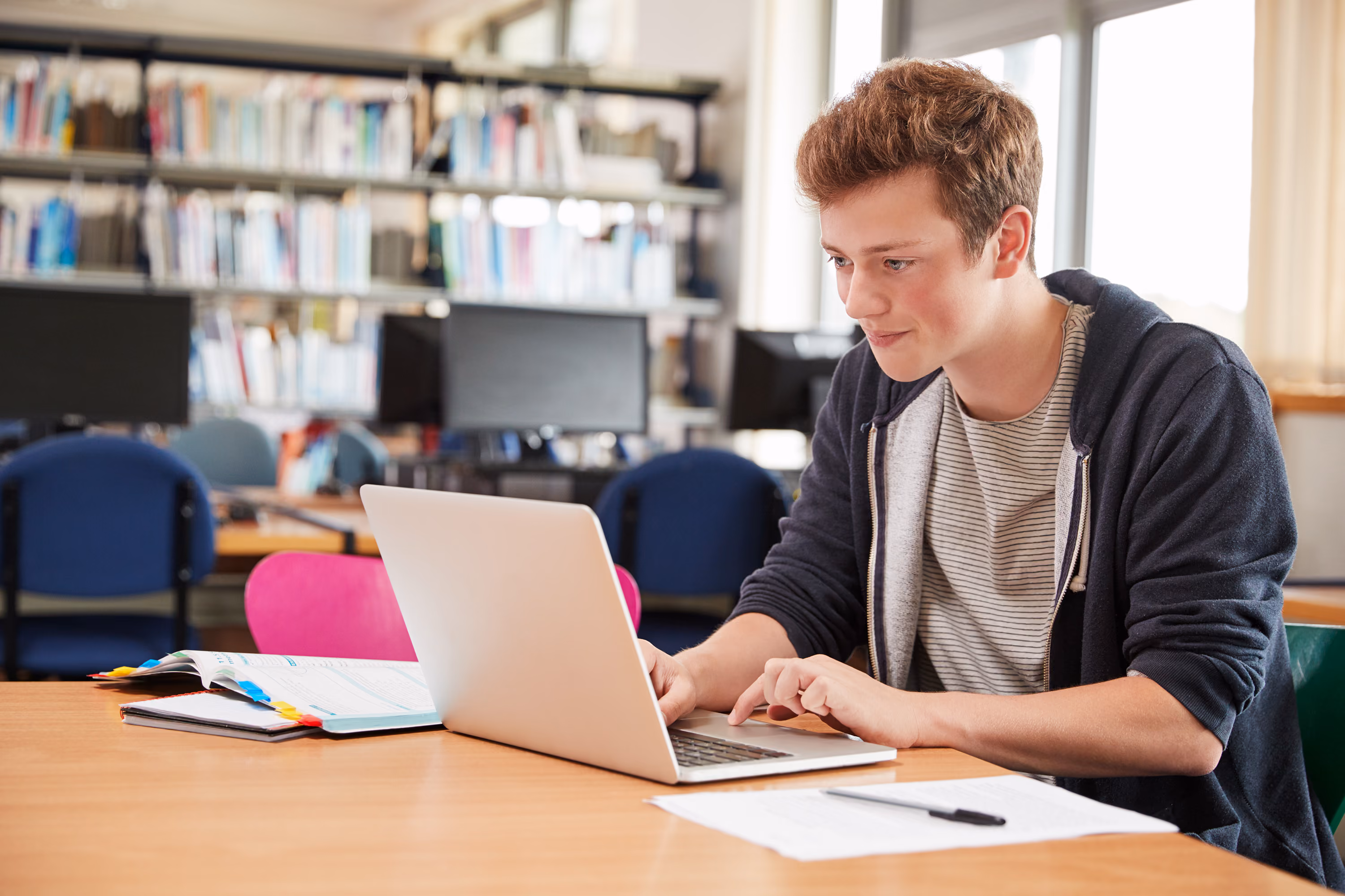 Young man working on a laptop in a library with books and computer screens in the background.