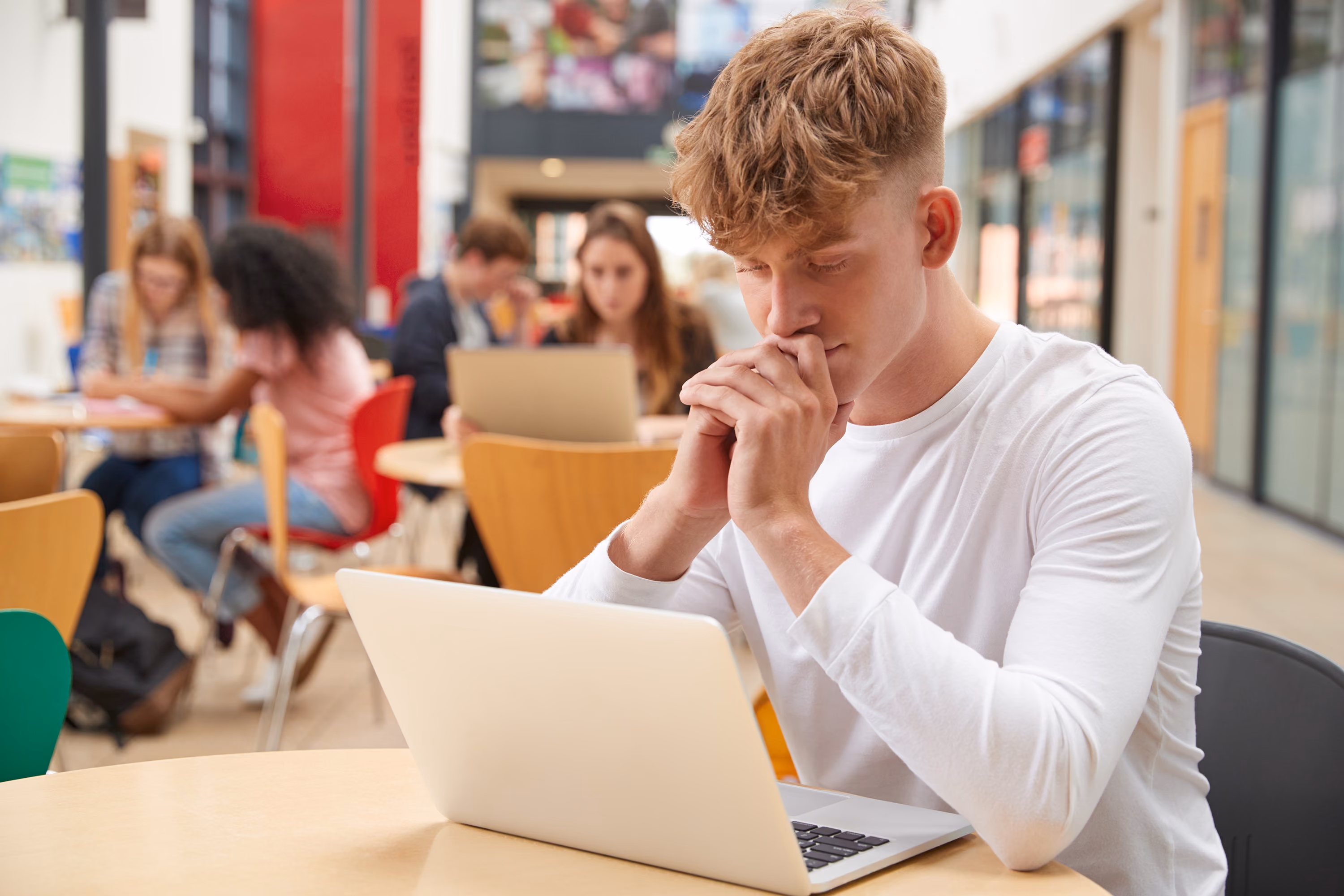 Teenage boy in white shirt looking stressed while using a laptop at a table in a busy school common area.