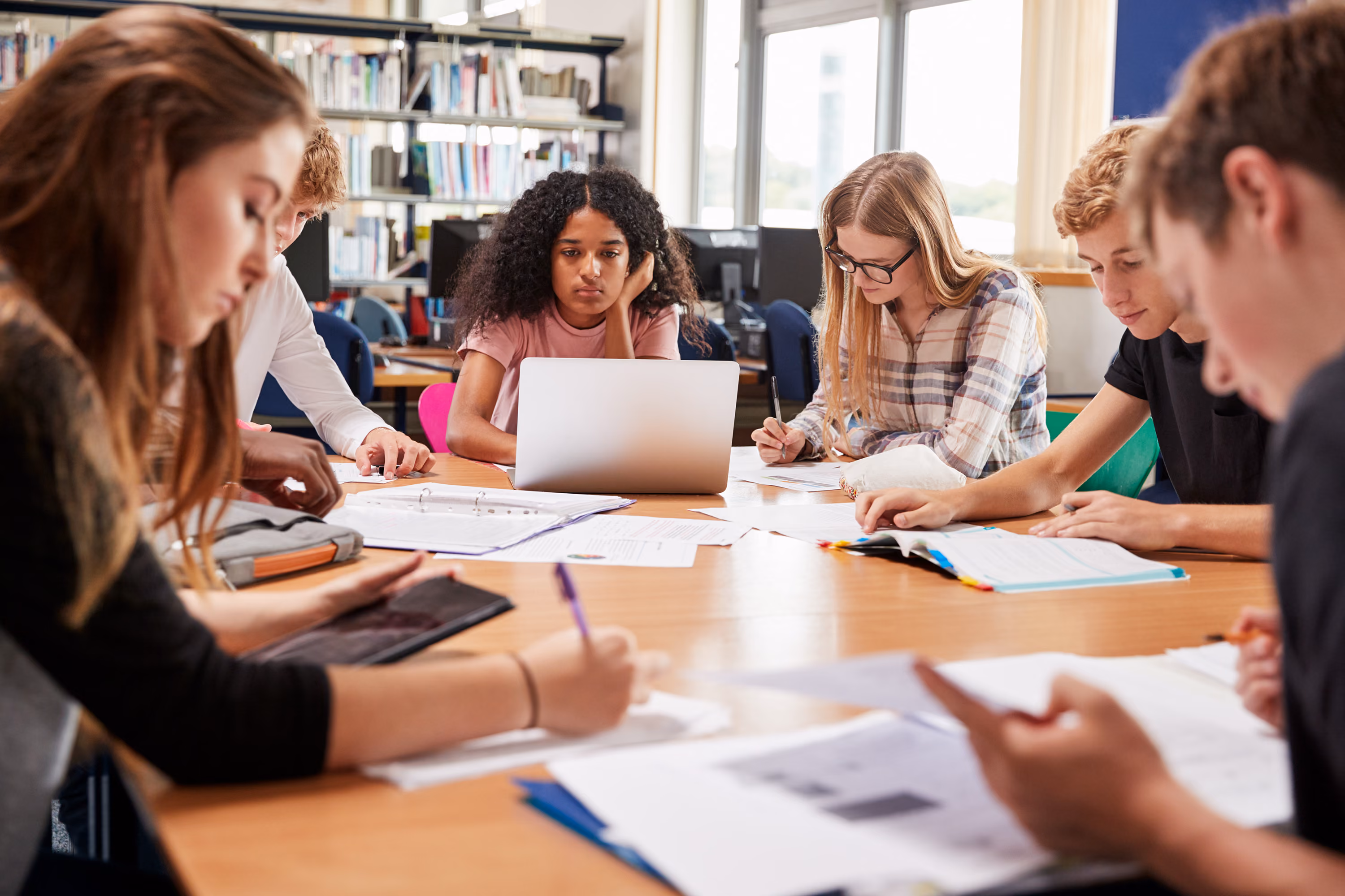 A group of teens studying around a table with books, papers, and a laptop in a classroom.