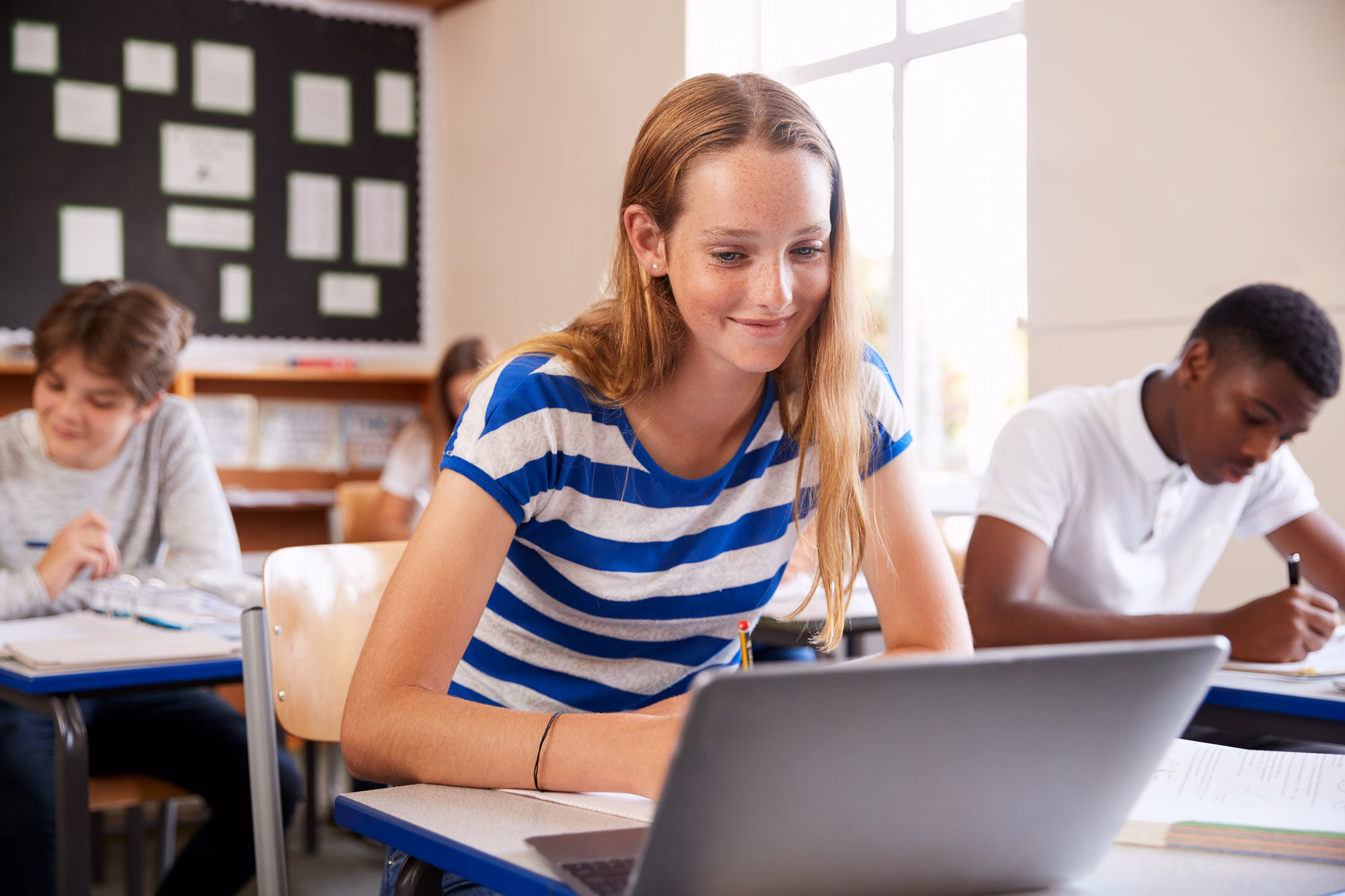 Teenage students studying in classroom, girl in striped shirt working on laptop at front desk.