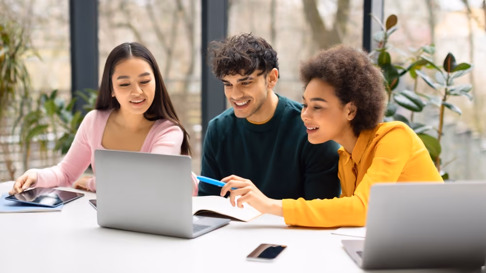 Three young people sitting at a table looking at a laptop and discussing with a tablet and phone nearby.