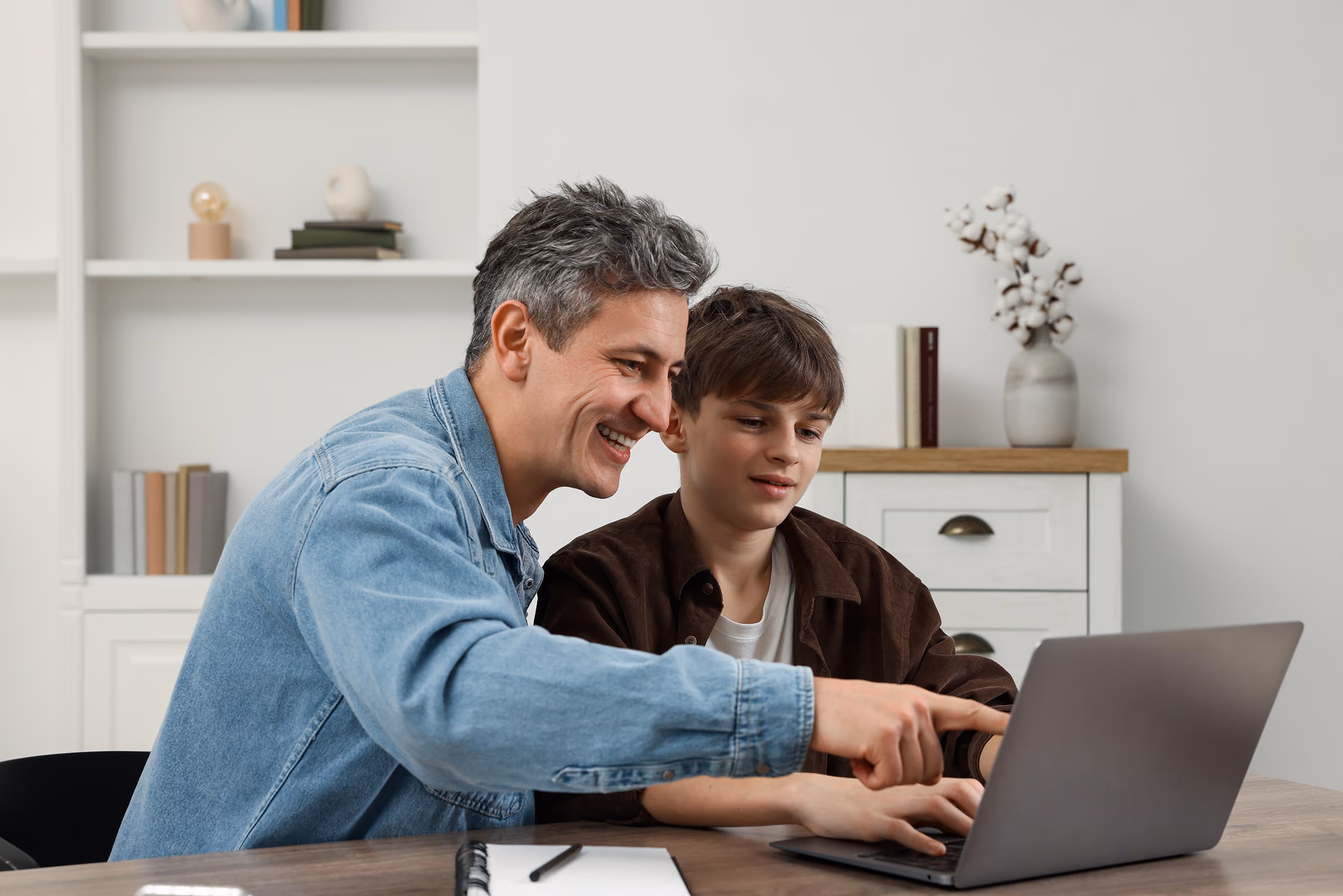 Smiling man in denim jacket teaching a boy sitting at a table with a laptop.