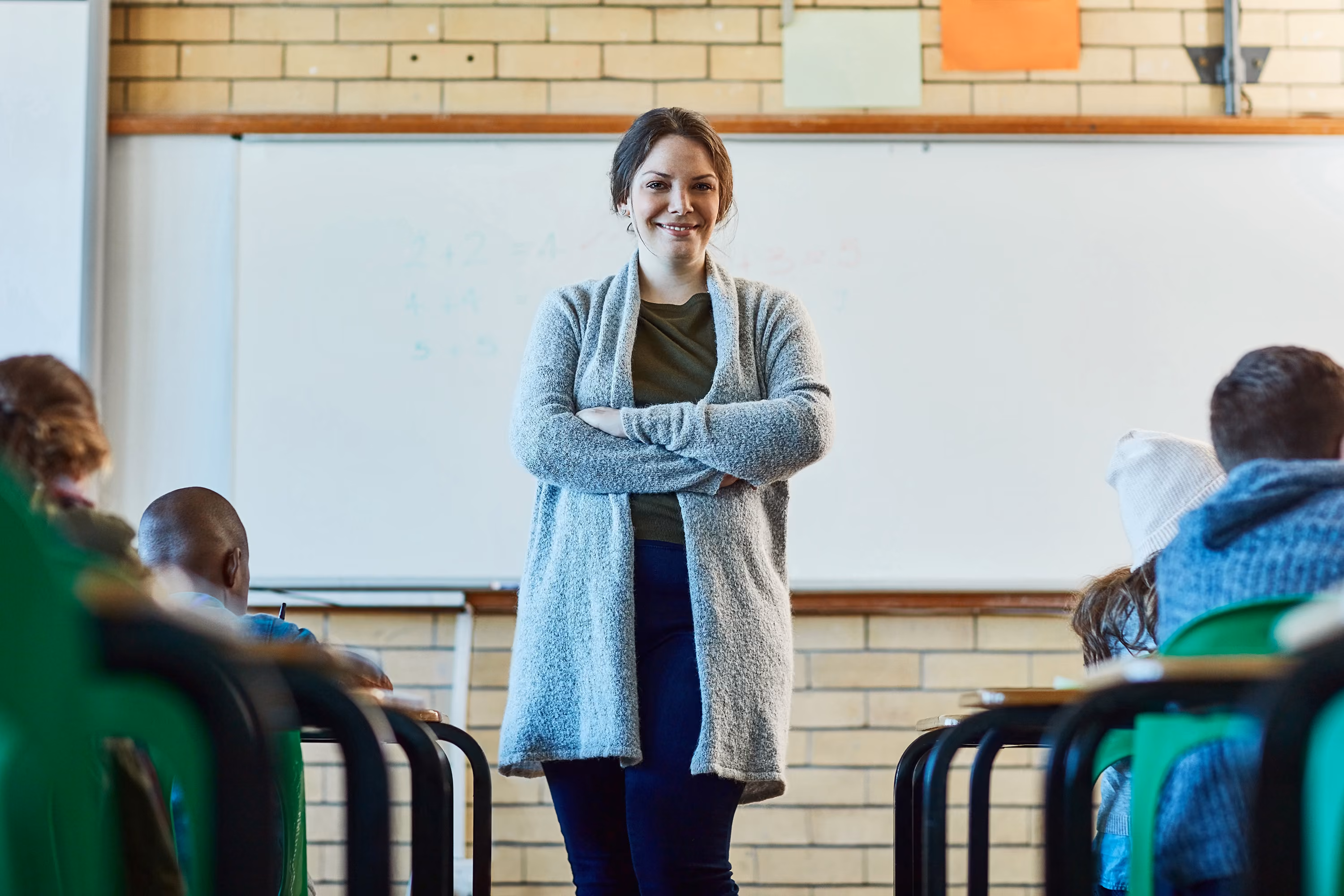 Smiling female teacher standing with arms crossed in front of whiteboard in classroom with seated students.