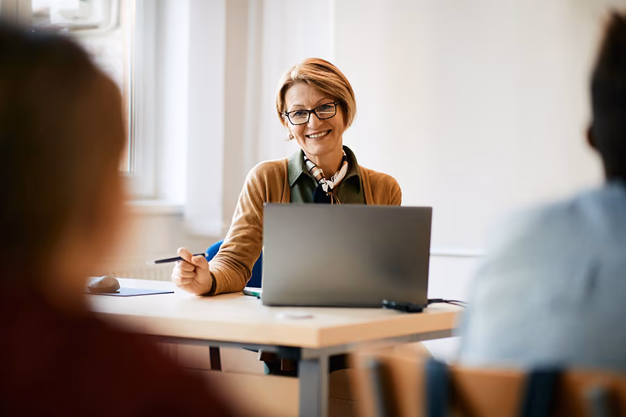 Smiling woman with glasses sitting at a desk with a laptop, engaging with two people.