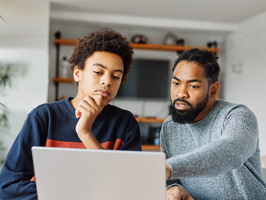 Man with beard teaching teenage boy using a laptop in a home setting with shelves in background.