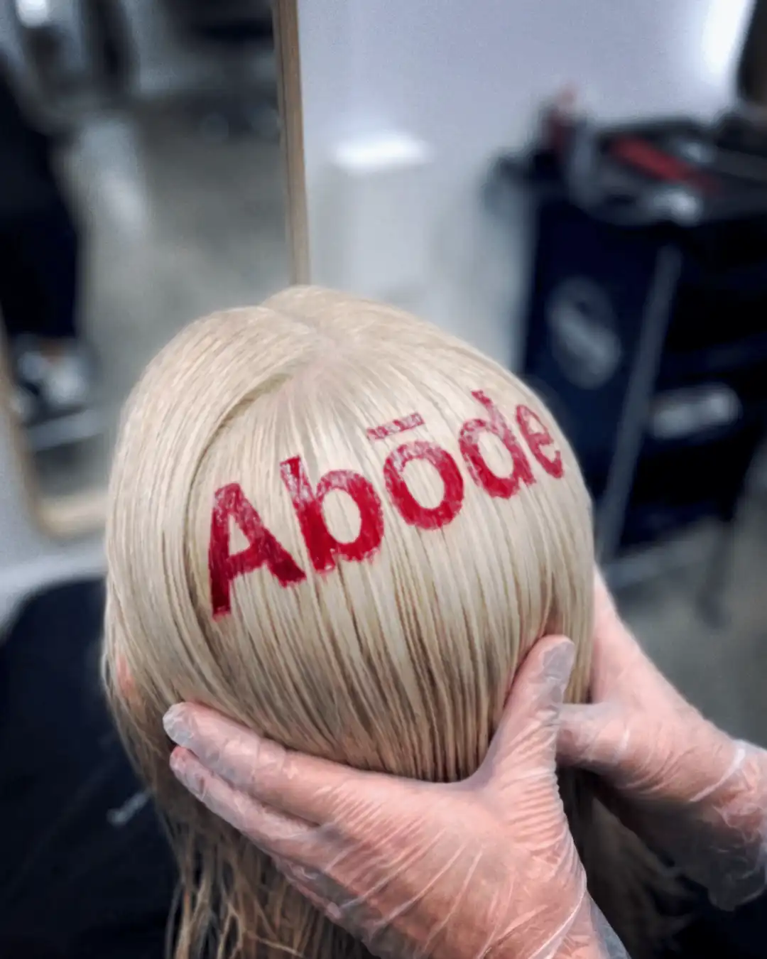 Gloved hands cradling freshly lightened platinum blonde hair, with Abōde hand-painted across it in red colour.
