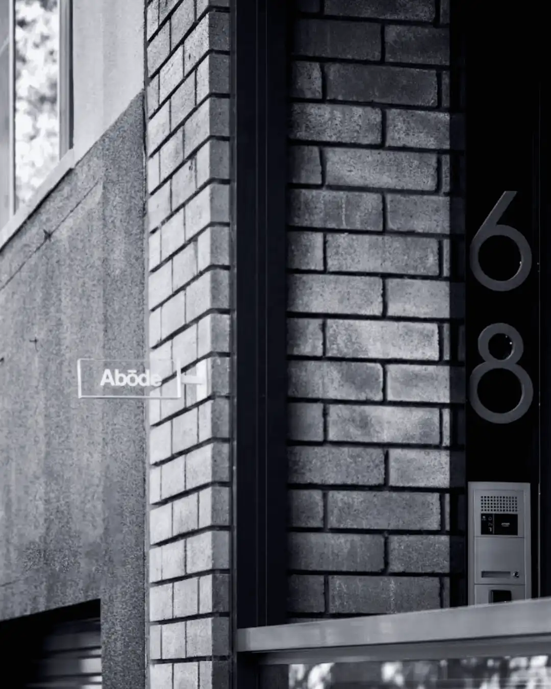 Black and white photo of the Abōde Hair Studio entrance, a small glass sign on brickwork beside the number 68.