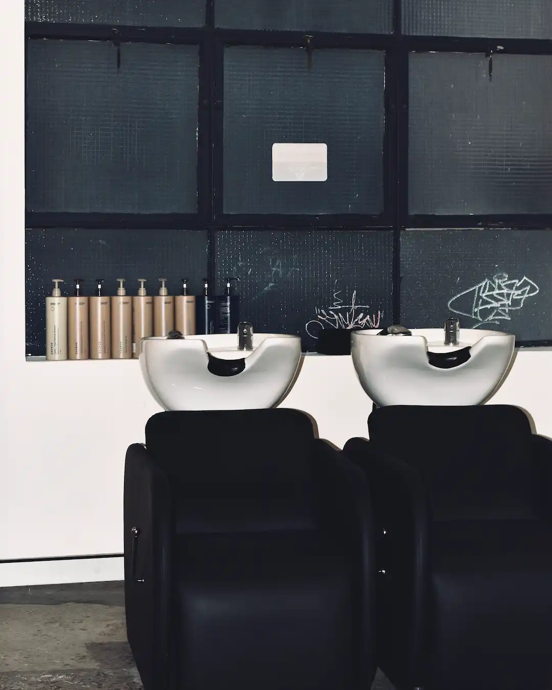 Two white backwash basins with black reclining chairs against a dark glass wall lined with Abōde haircare products.