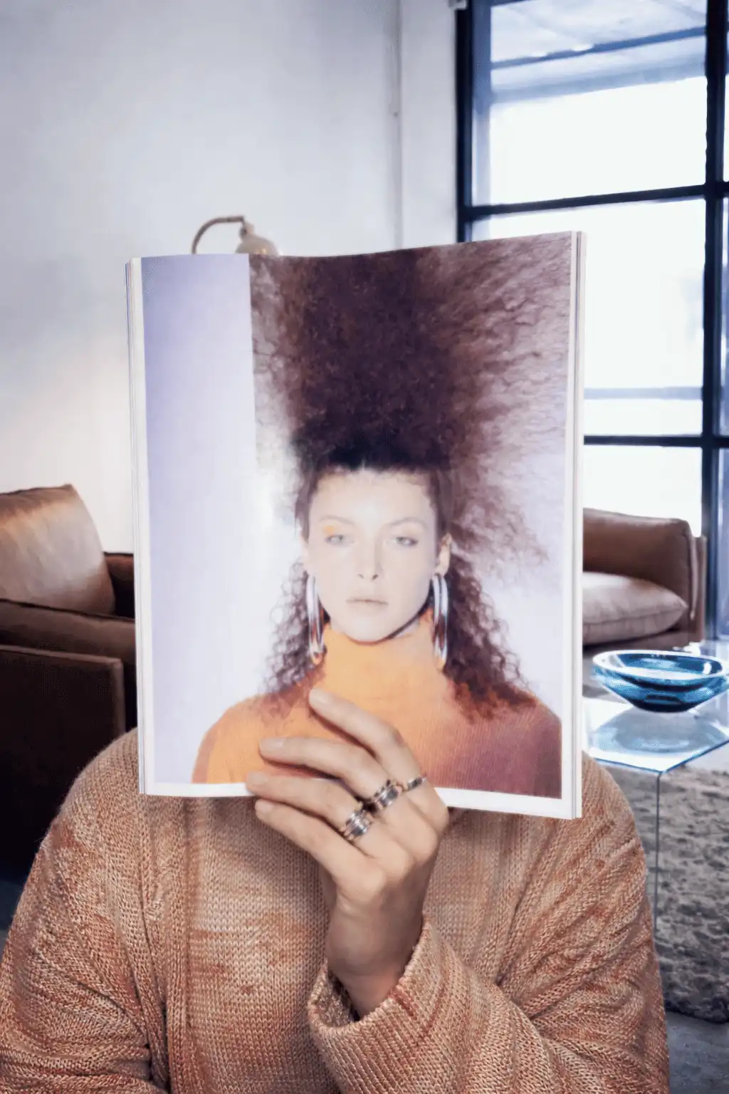Person in a knit sweater holding an open magazine over their face, showing a model with voluminous curls and hoop earrings.