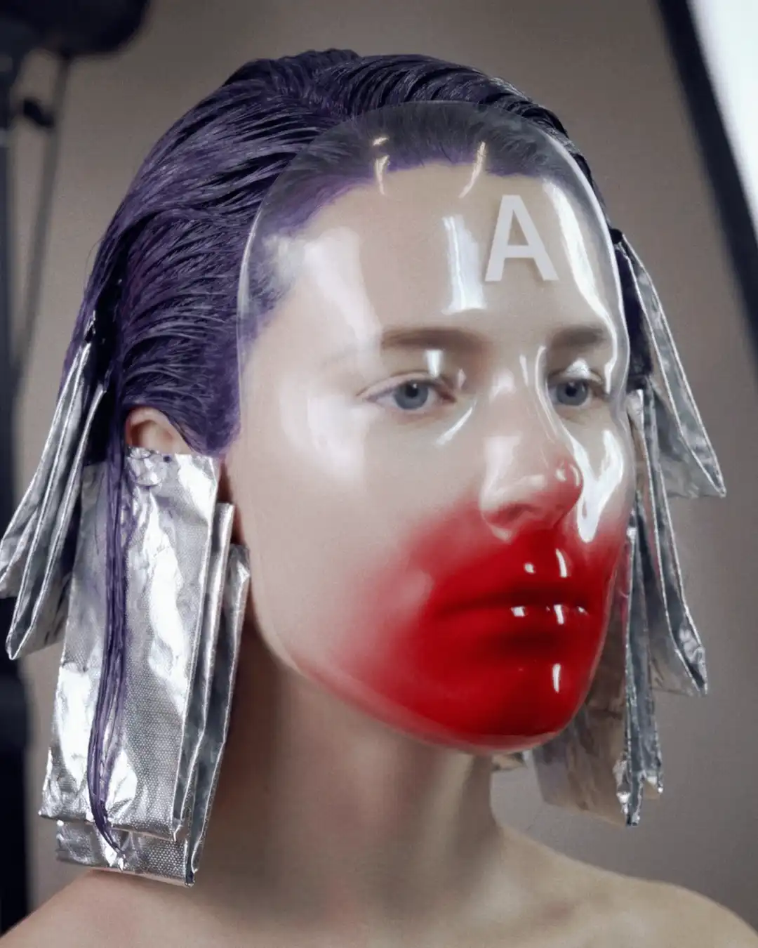 Model with purple-toned wet hair and silver foils, wearing a clear moulded mask washed with red at the mouth.