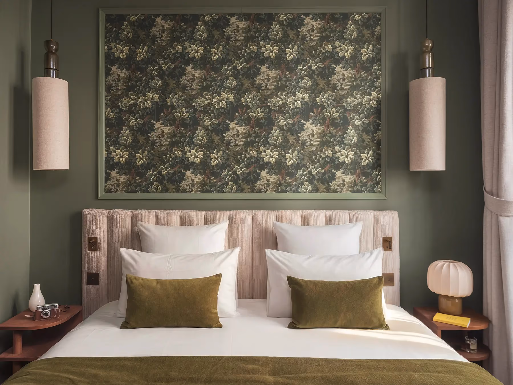 Tête de lit capitonnée beige encadrée de deux appliques, tapisserie florale verte et coussins olive.