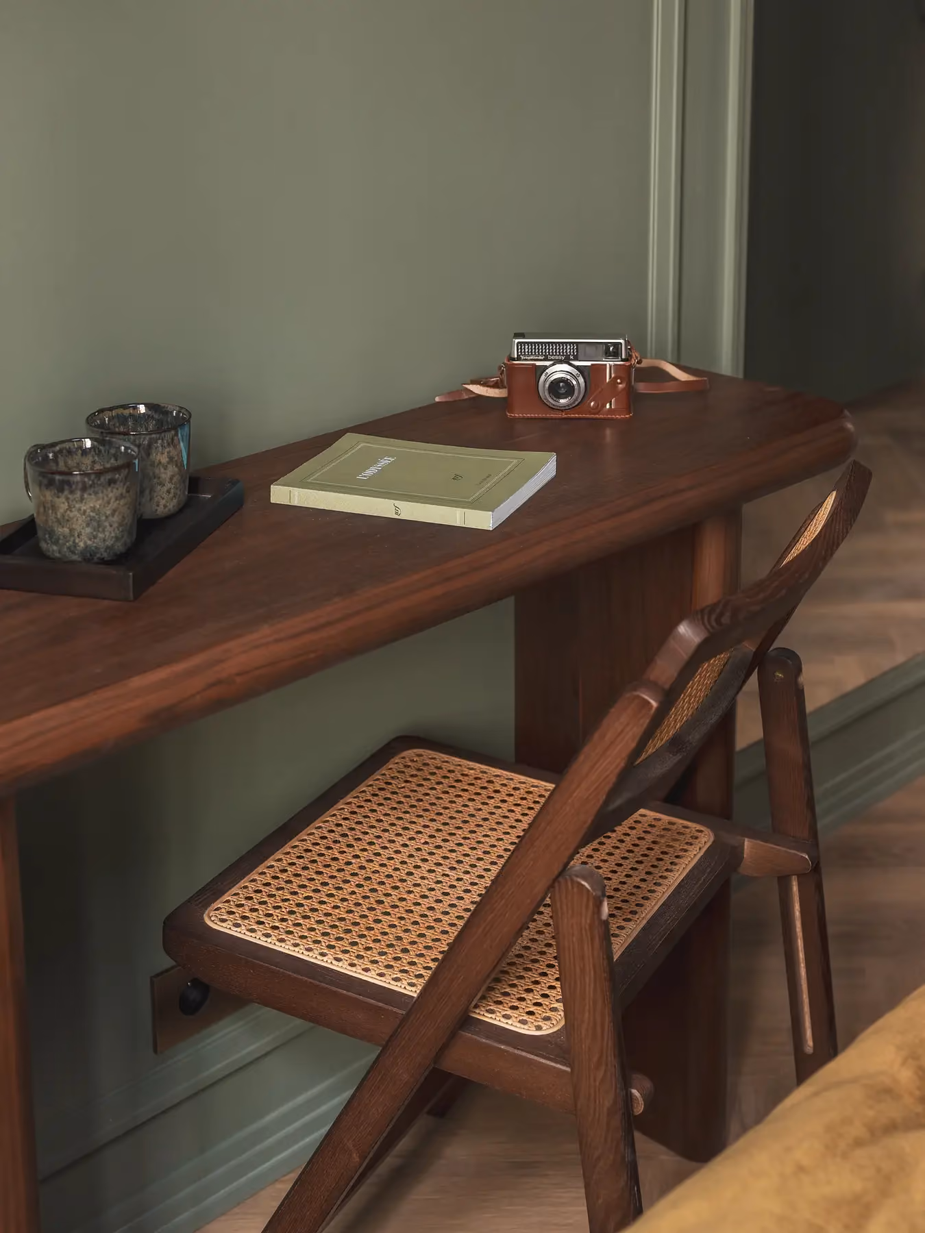 Bureau en bois avec appareil photo vintage, livre et chaise pliante cannée, mur vert.